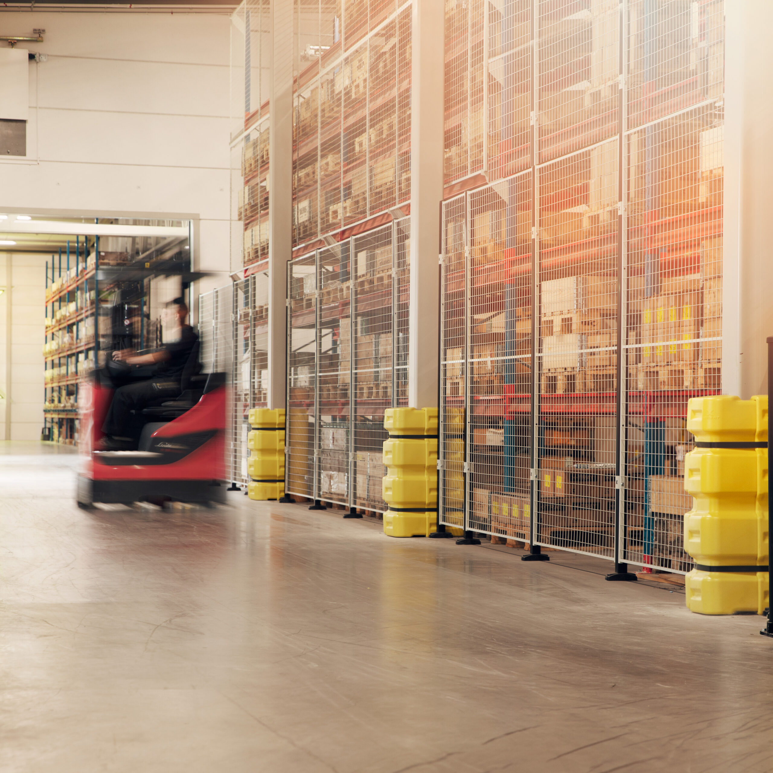 Rode heftruck rijdt voorbij een rij geel-zwarte kolombeschermers langs magazijnstellingen in een grote industriële opslagruimte.
