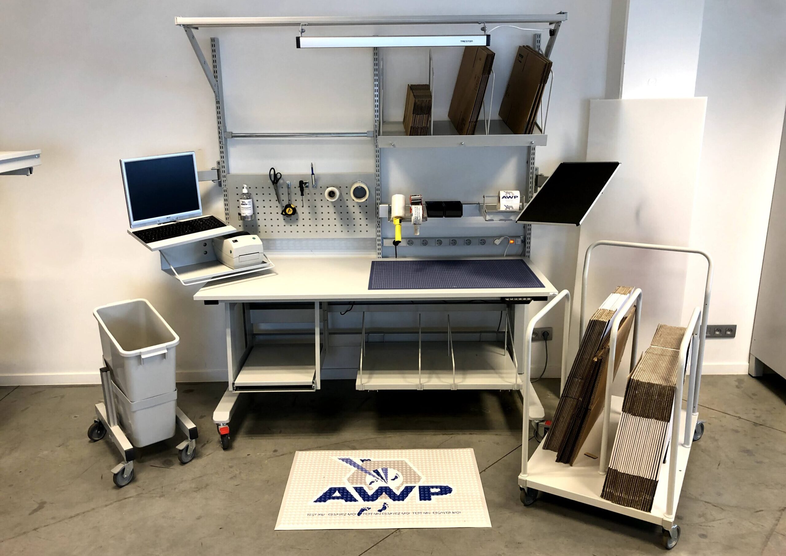 Volledig uitgerust AWP inpakwerkstation met monitor, toetsenbord, printer, gereedschapsbord, tape-dispenser, inpakmateriaal en een ergonomische antivermoeidheidsmat op de vloer.