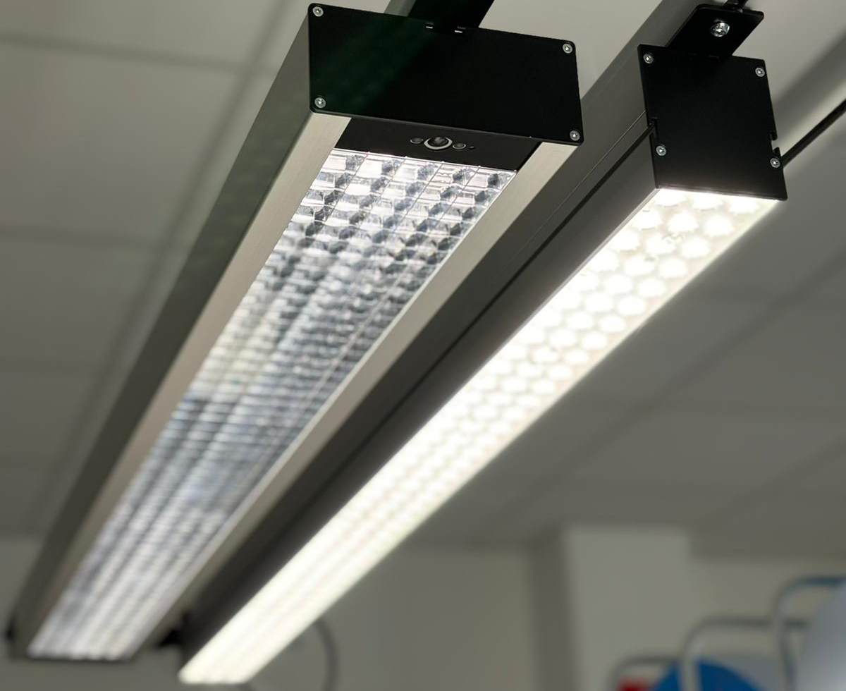 Twee lineaire LED-verlichtingsarmaturen gemonteerd aan het plafond boven een werkplek, met schakelaar en verstelbare beugels zichtbaar.
