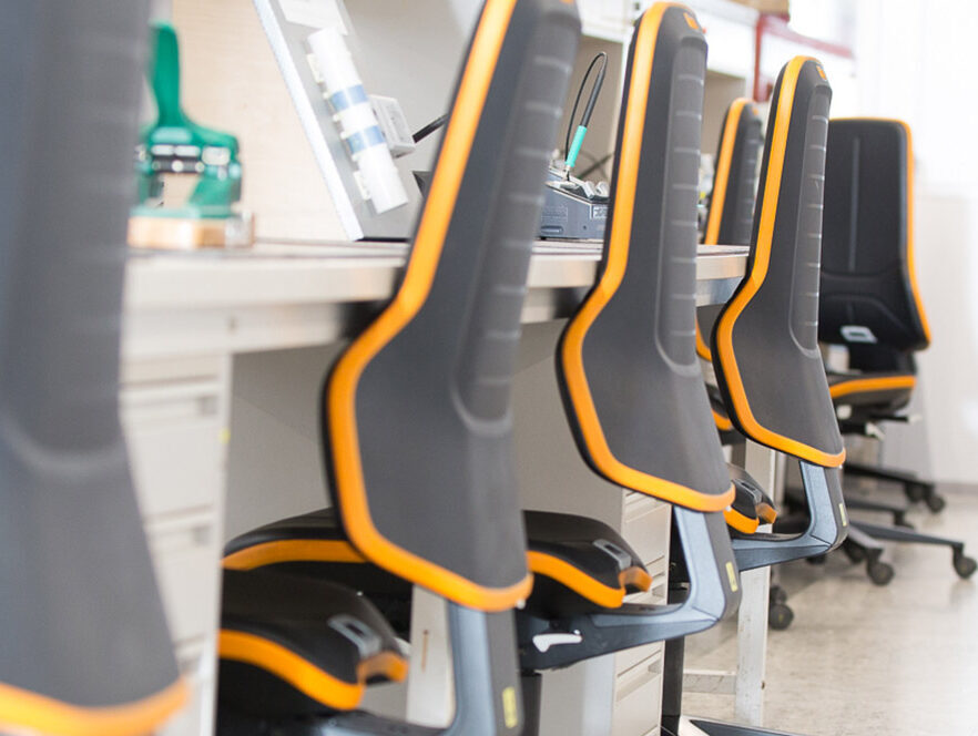 Rij zwarte ergonomische industriële werkstoelen bimos, opgesteld aan grijze werktafels in een productieomgeving.