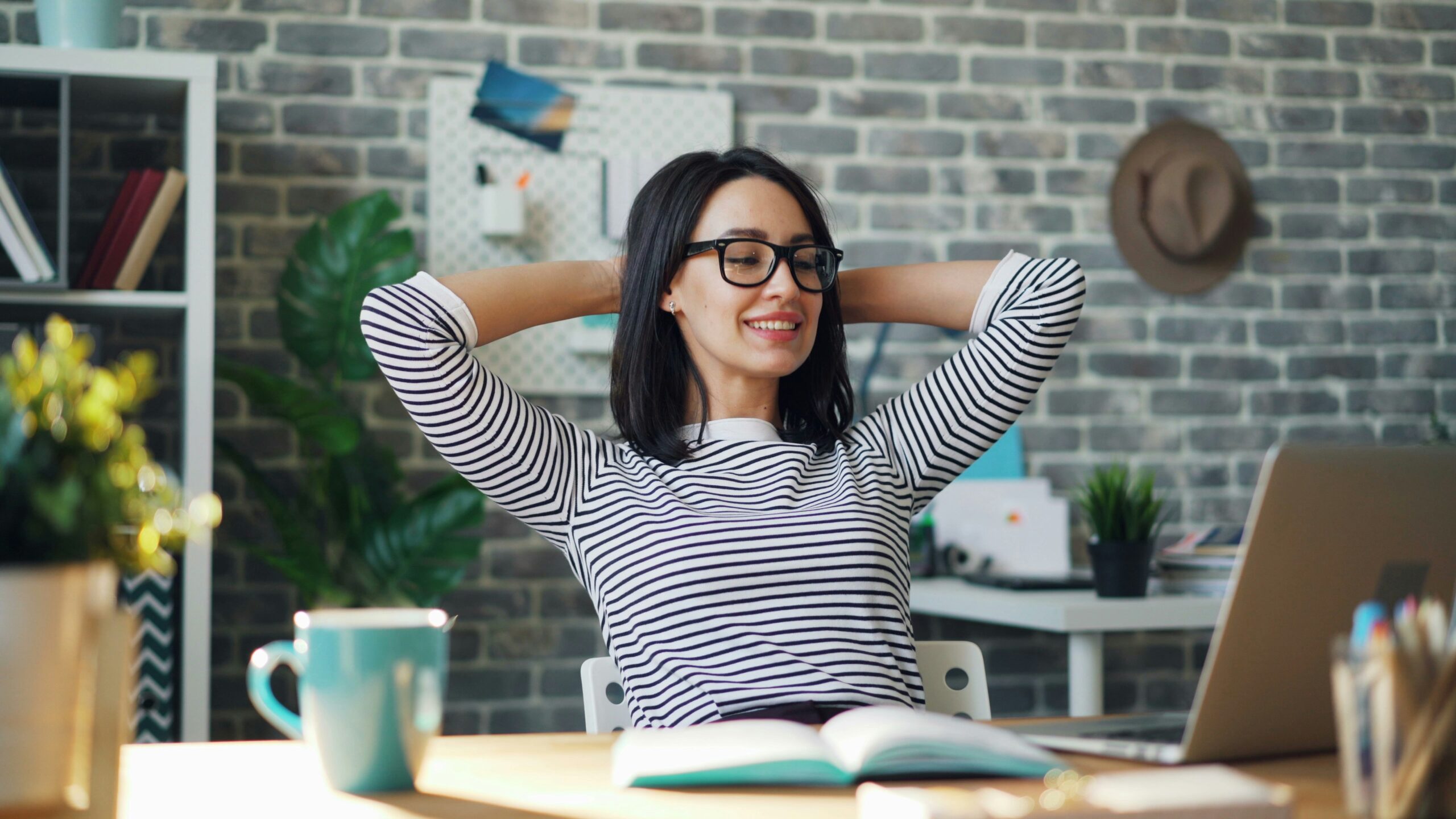 Lachende vrouw met bril legt haar handen achter haar hoofd en strekt haar rug aan een houten bureau met laptop en notitieboek, in een gezellige kantooromgeving.