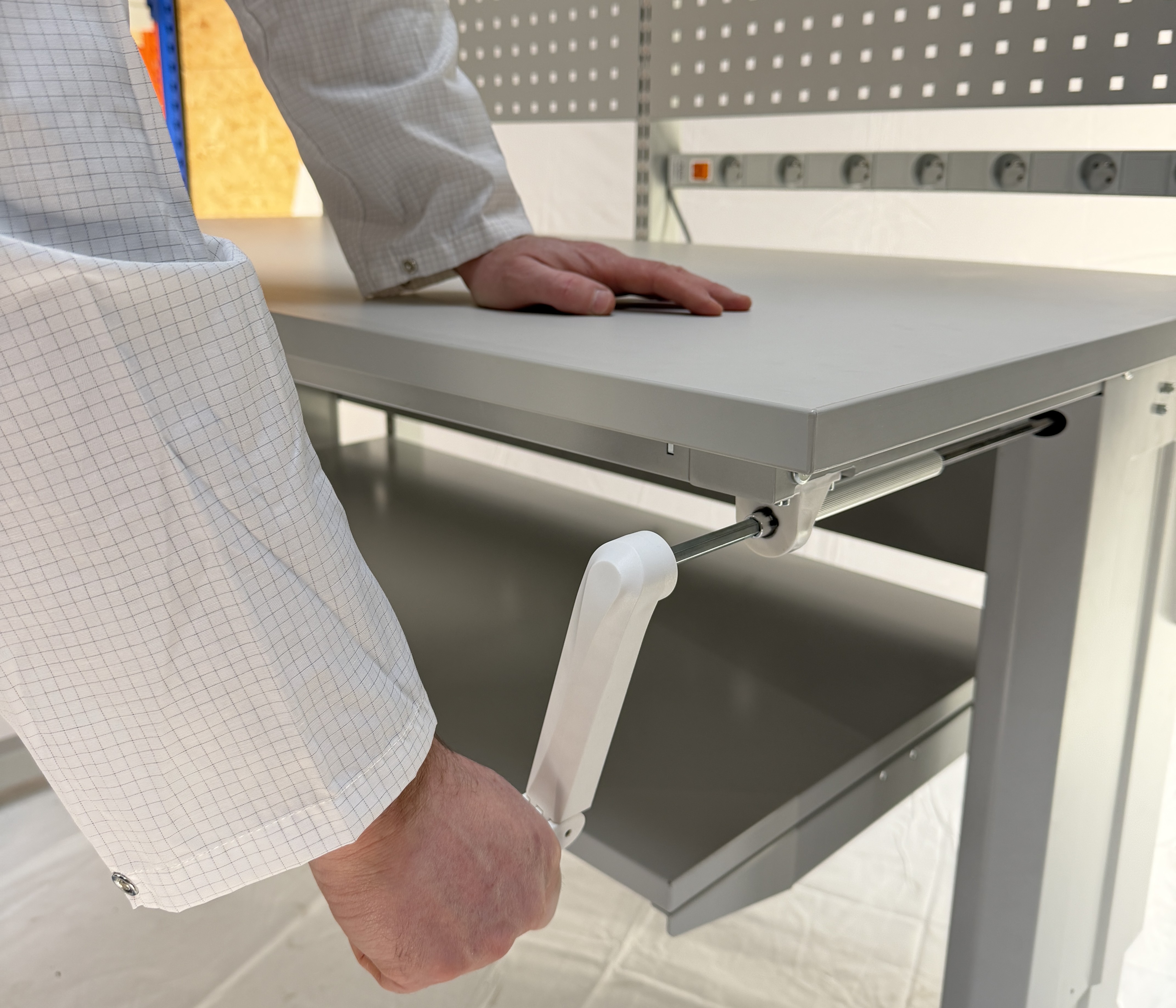 Hand draait een witte handzwengel aan de zijkant van een grijze in hoogte verstelbare werktafel in een industriële werkruimte.