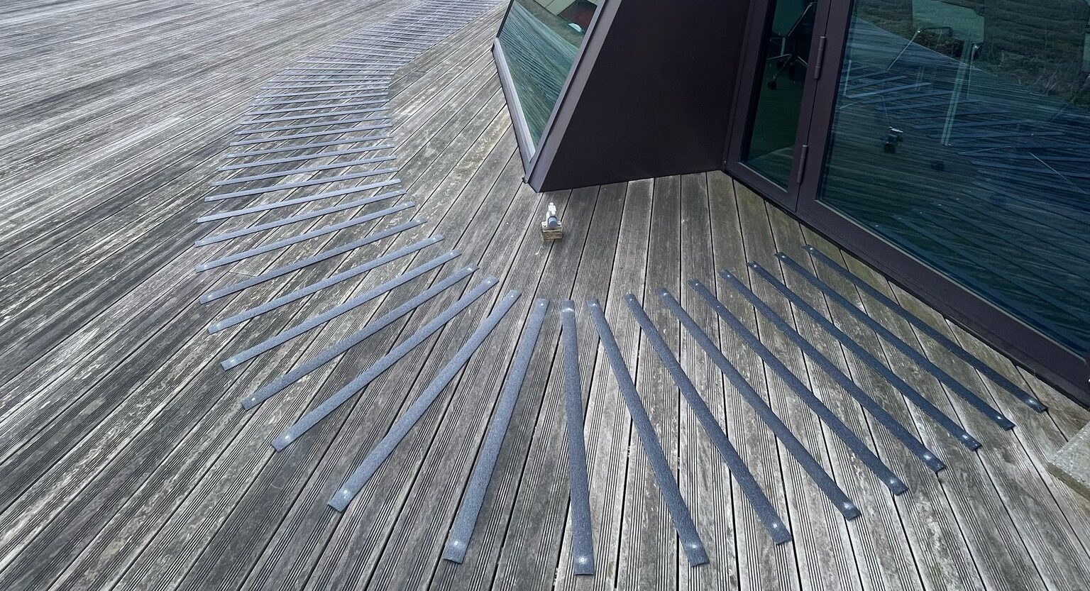 Grijze GRP antislip stroken diagonaal aangebracht op een verweerd houten terras naast een modern gebouw met glazen geveldelen