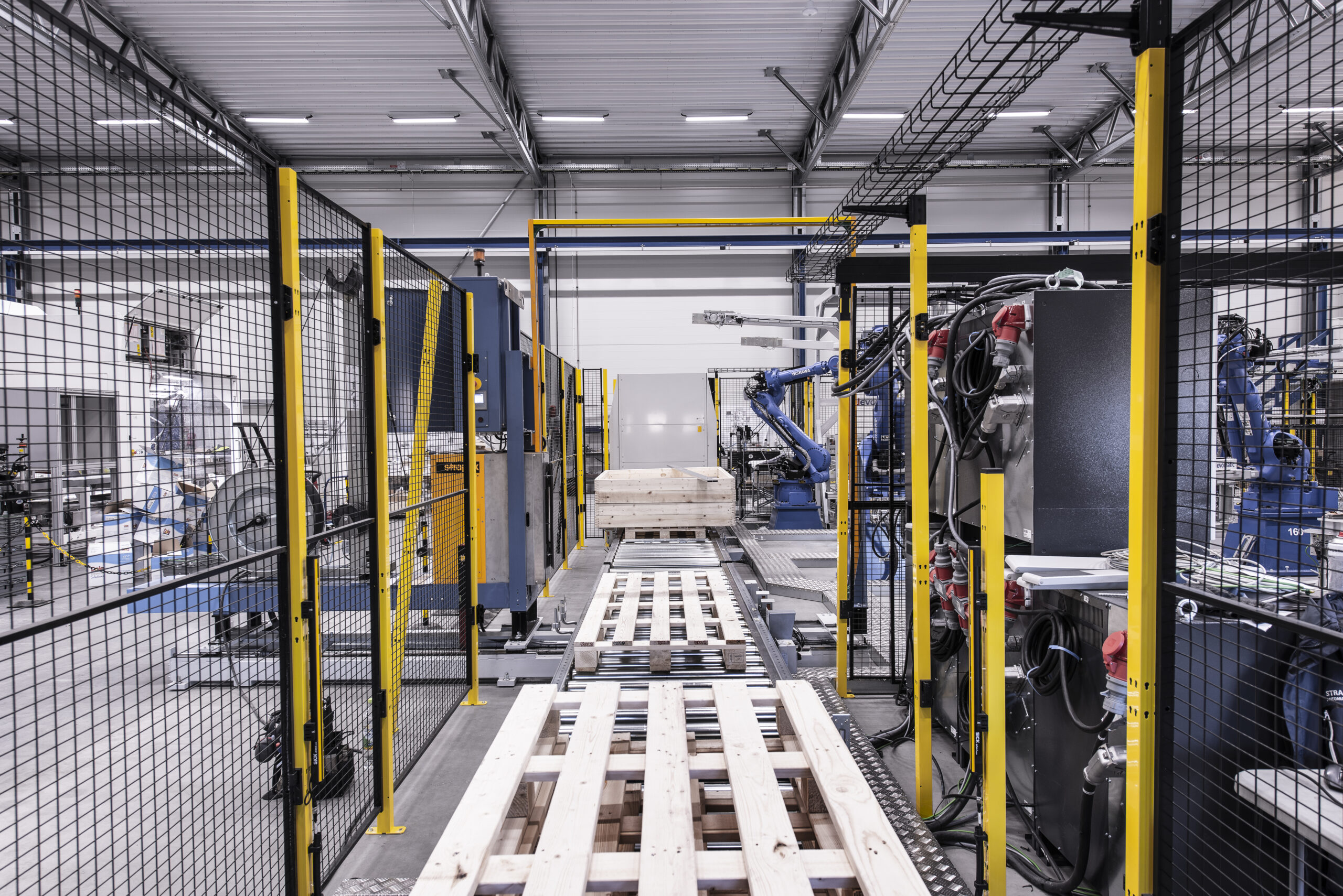 Geautomatiseerde productielijn afgeschermd met zwarte gaaswanden op gele stalen palen, met robotarmen en een palletconveyor zichtbaar achter het hekwerk.