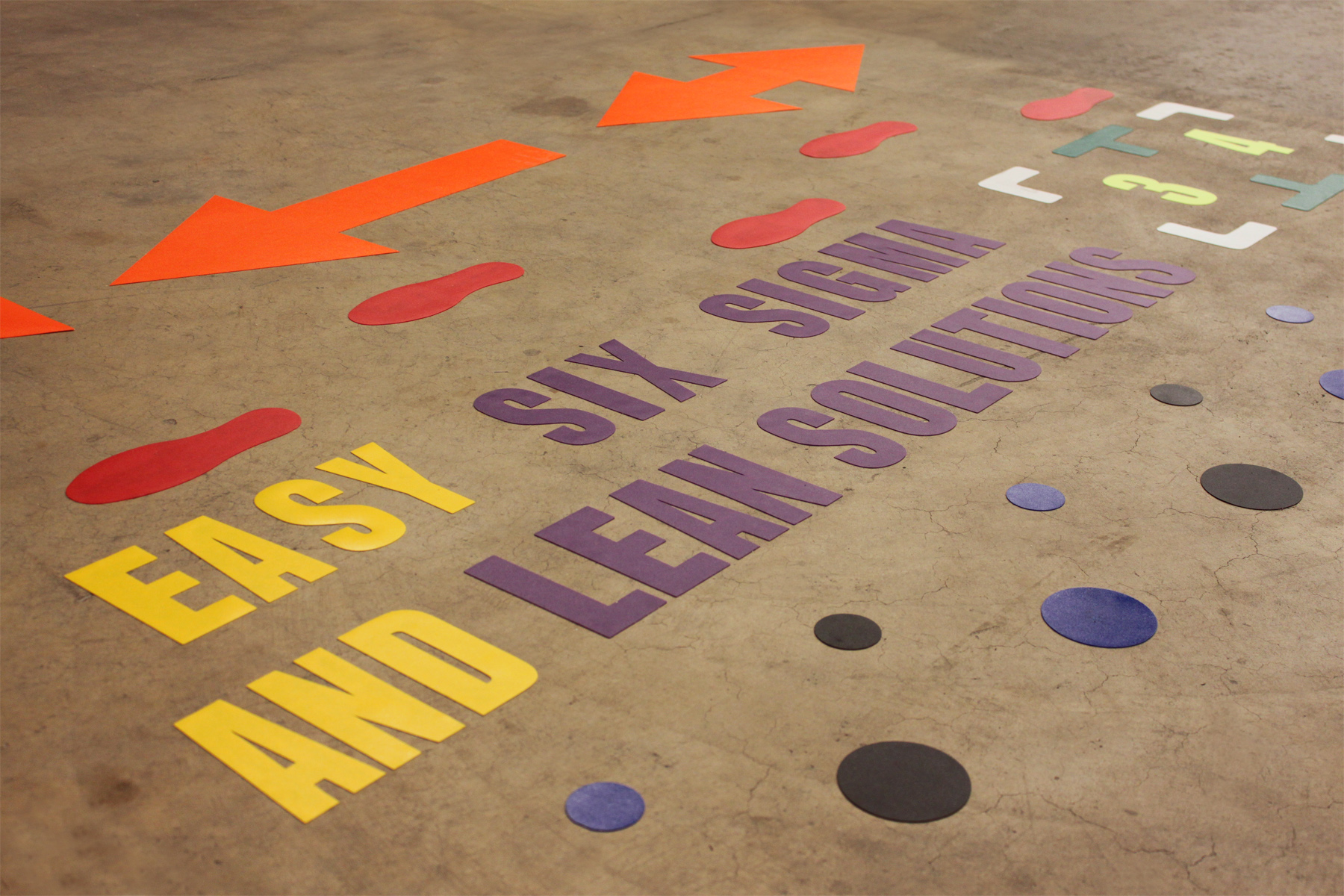 Betonnen industriële vloer met kleurrijke vloerstickers: oranje pijlen, rode voetafdrukken, gekleurde stippen, cijfers en de tekst "Easy Six Sigma and Lean Solutions"