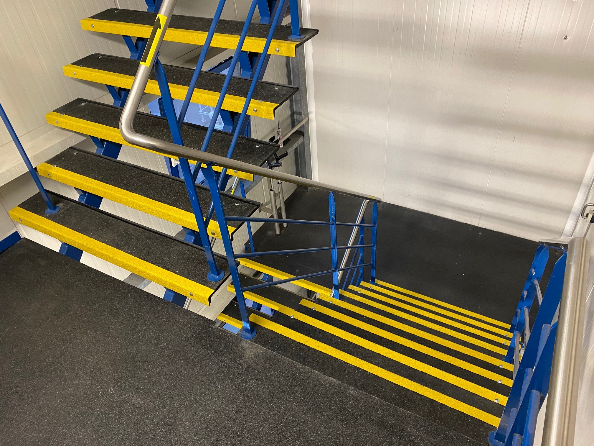 Zwarte rubber trappen met gele antislip trapprofielen en blauwe stalen leuningen in een witte industriële ruimte, vermoedelijk in de voedingsindustrie / Escaliers en caoutchouc noir avec profilés de marche antidérapants jaunes et rampes en acier bleu dans un espace industriel blanc, probablement dans l'industrie alimentaire