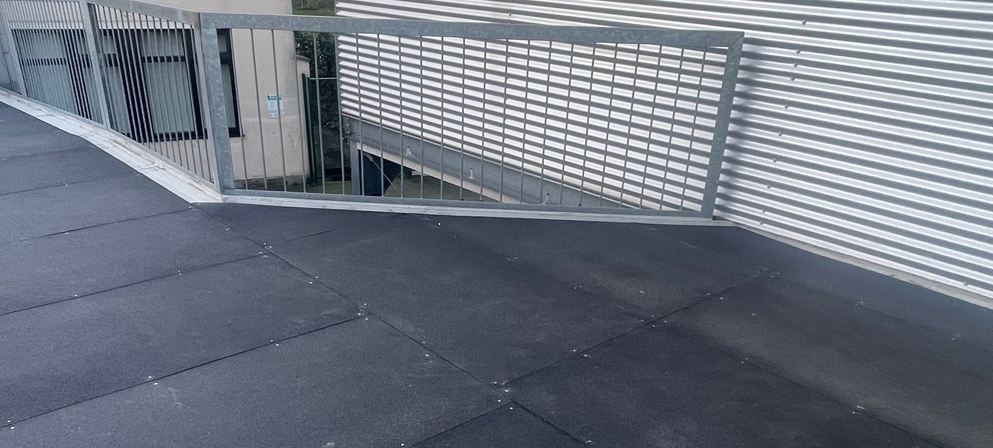 Donkergrijze GRP antislip tegels aangebracht op een plat dak, met een stalen hekwerk en golfplaten gevel op de achtergrond