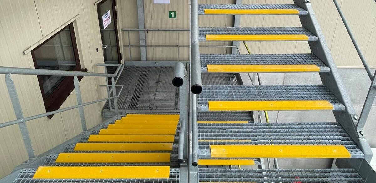 Dubbele buitentrap met stalen roostervloer en gele antislip trapprofielen op elke trede, naast een industrieel gebouw