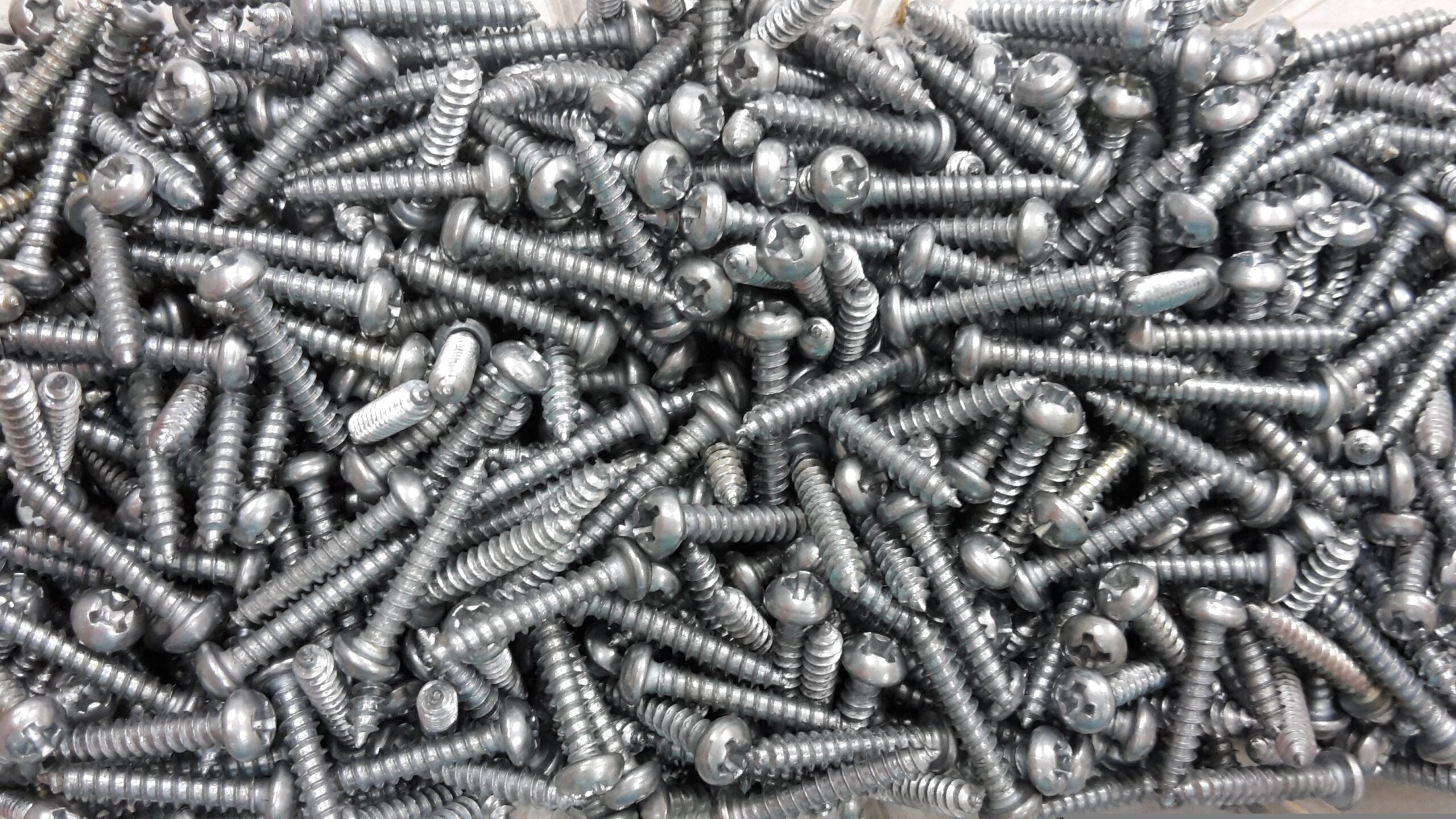 Close-up van een grote hoeveelheid grijze metalen schroeven met kruiskop, los gestort op een hoop.