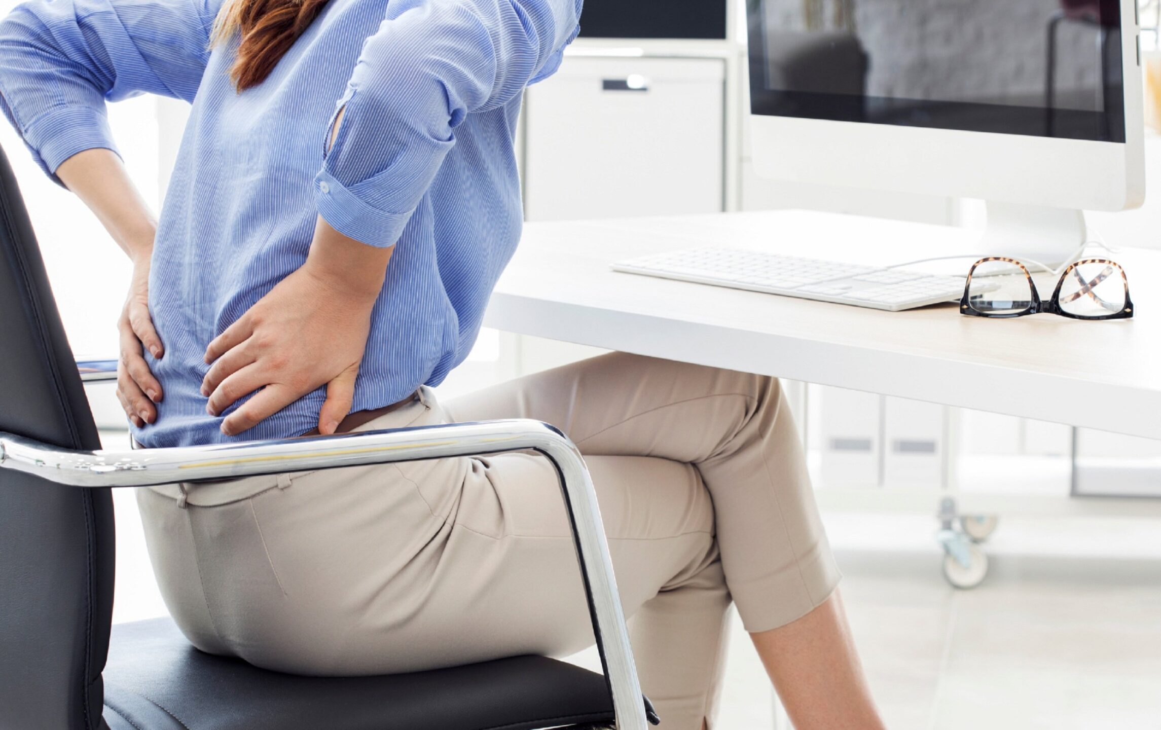 Vrouw in lichtblauwe blouse houdt haar onderrug vast terwijl ze op het puntje van haar stoel zit aan een bureau, met een bril en laptop op de achtergrond.