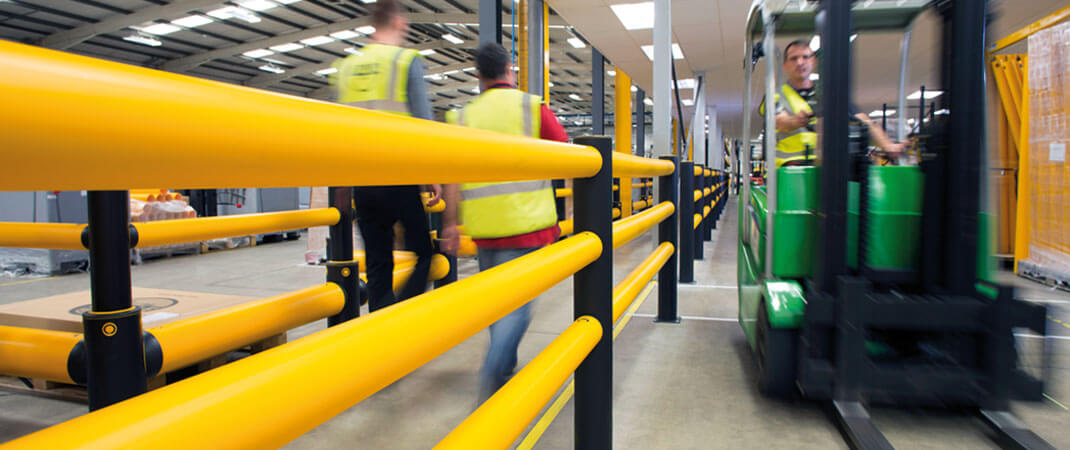 Medewerkers in veiligheidshesjes lopen langs een gele stalen veiligheidsrailing in een drukke industriële of logistieke ruimte / Des employés en gilets de sécurité marchent le long d'une rambarde de sécurité en acier jaune dans un espace industriel ou logistique animé