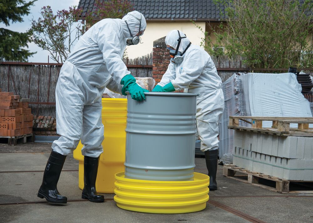 Twee medewerkers in witte beschermingspakken, gasmaskers en groene handschoenen hanteren een grijze drum op een gele opvangbak buiten.