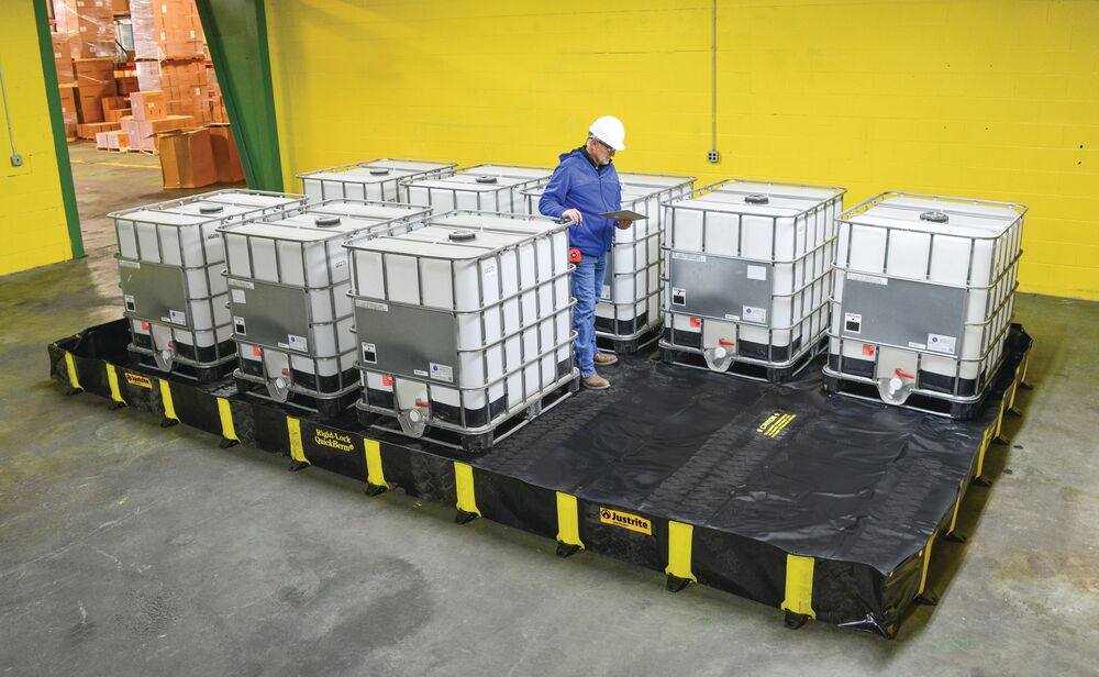 Medewerker met helm die IBC-containers controleert op een grote zwarte flexibele Justrite opvangbak in een industrieel magazijn