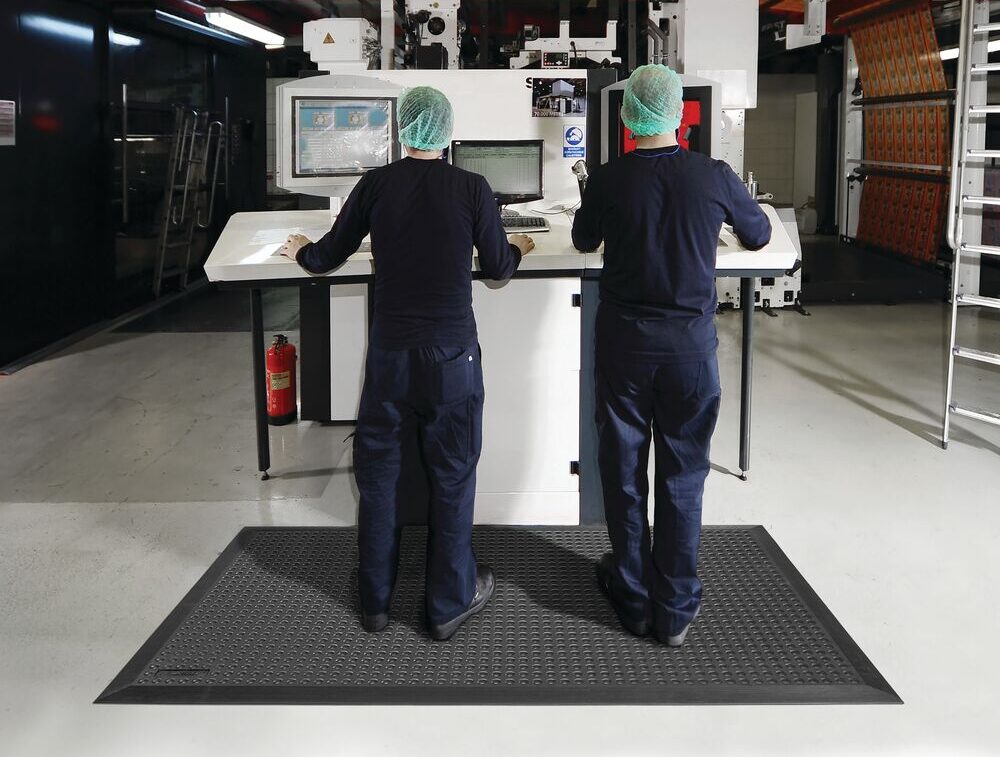 Twee medewerkers in donkerblauwe werkkleding en haarnetjes staan op een grote zwarte ergonomische mat voor een productiemachine met twee monitoren in een industriële hal.