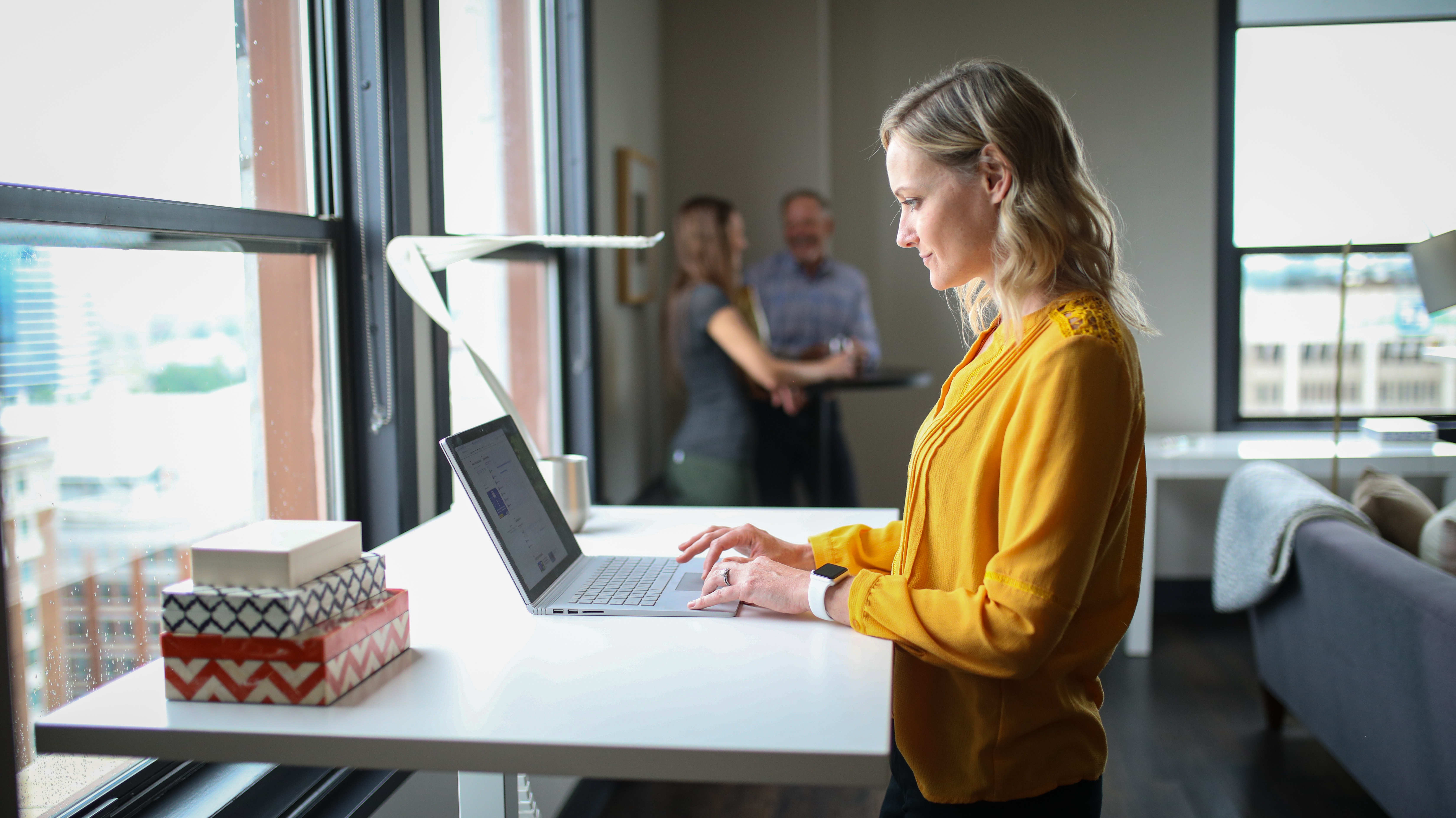 Vrouw in geel hemd typt staand op een laptop aan een wit in hoogte verstelbaar bureau bij een raam, met collega's op de achtergrond.