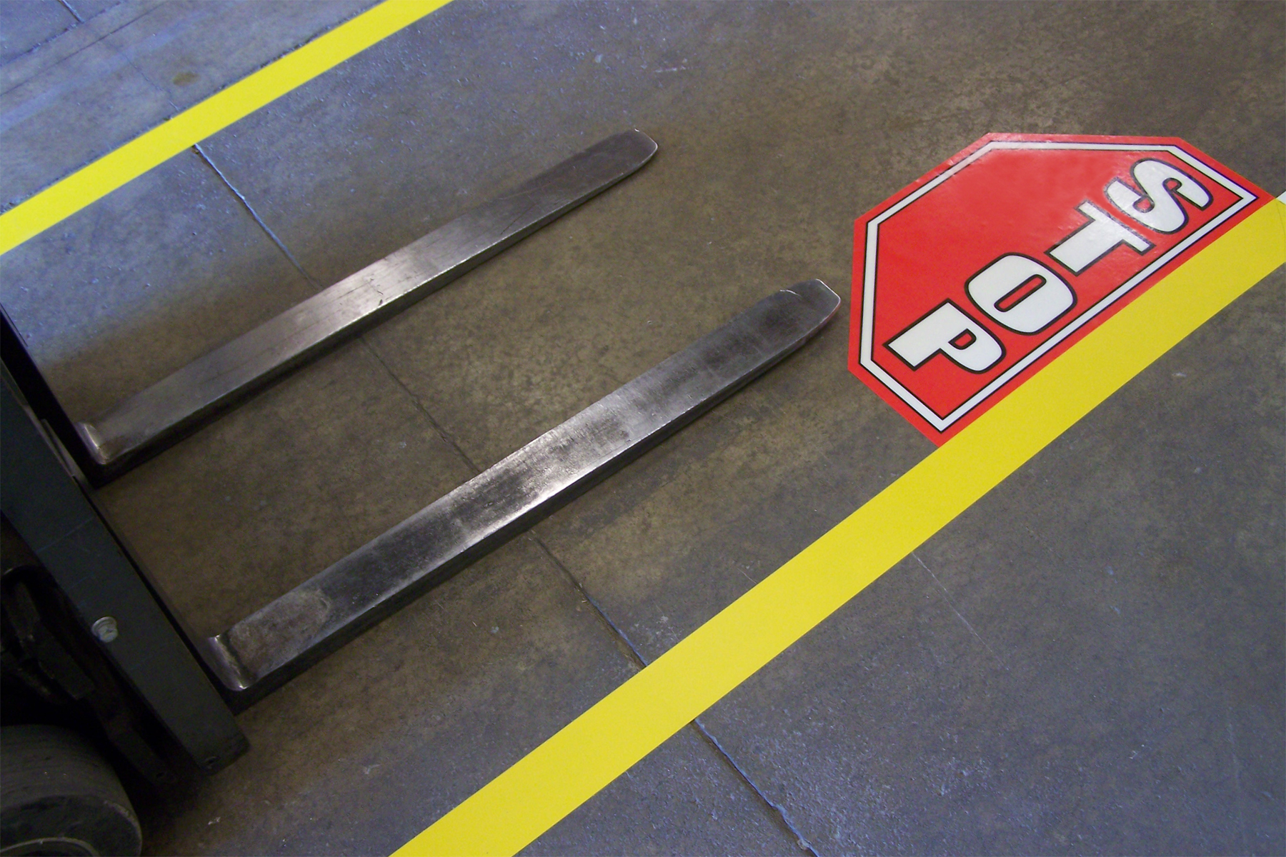 Vorken van een vorkheftruck naast een rood zeshoekig stopbord als vloersticker op een betonnen vloer met gele afbakeningslijn