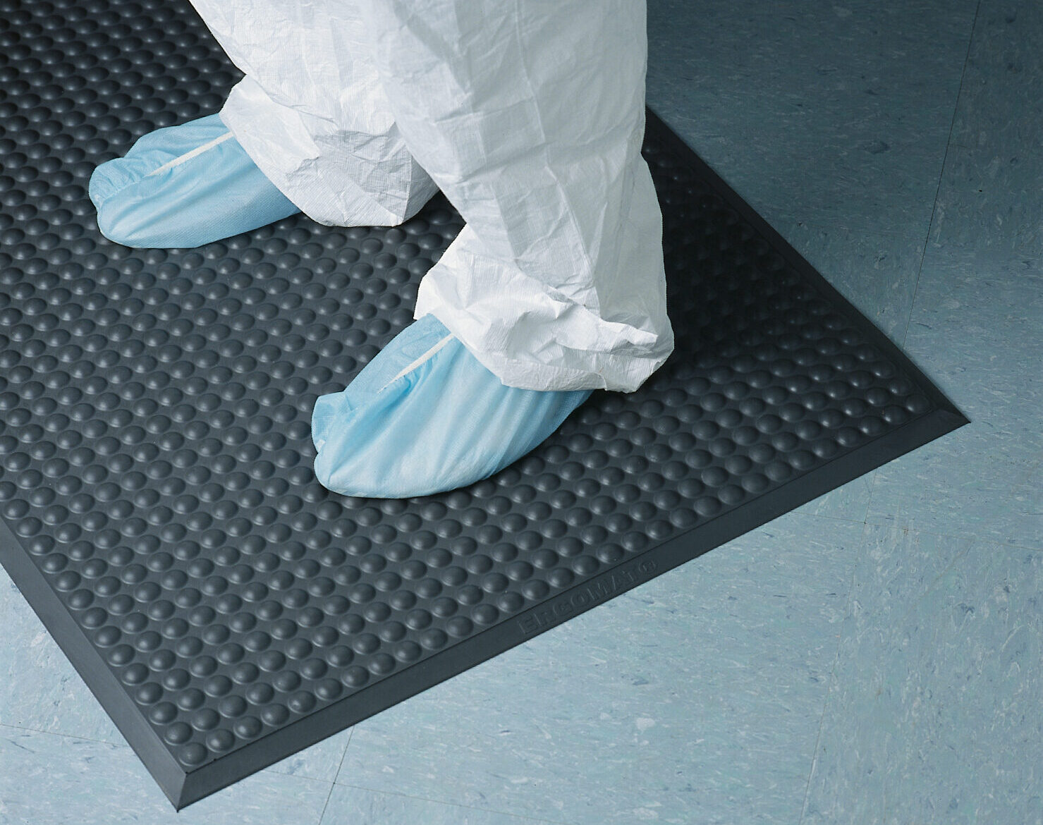 Close-up van voeten met blauwe wegwerpoverschoenen en witte beschermende overall, staand op een donkergrijze ergonomische stamat met noppenstructuur / Gros plan de pieds portant des sur-chaussures bleues jetables et une combinaison de protection blanche, debout sur un tapis anti-fatigue gris foncé à surface à picots