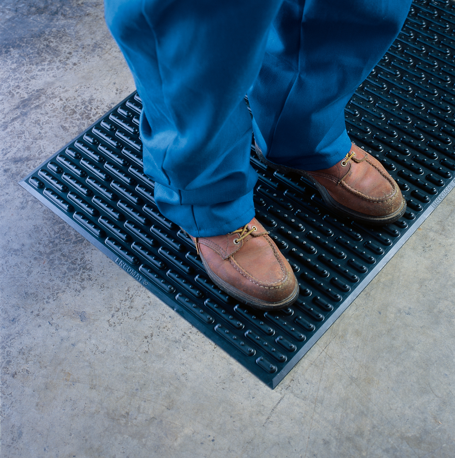 Onderbenen van persoon in blauwe werkbroek en bruine schoenen staand op een zwarte Ergomat Softline mat met langwerpig ribbelprofiel op een betonnen vloer