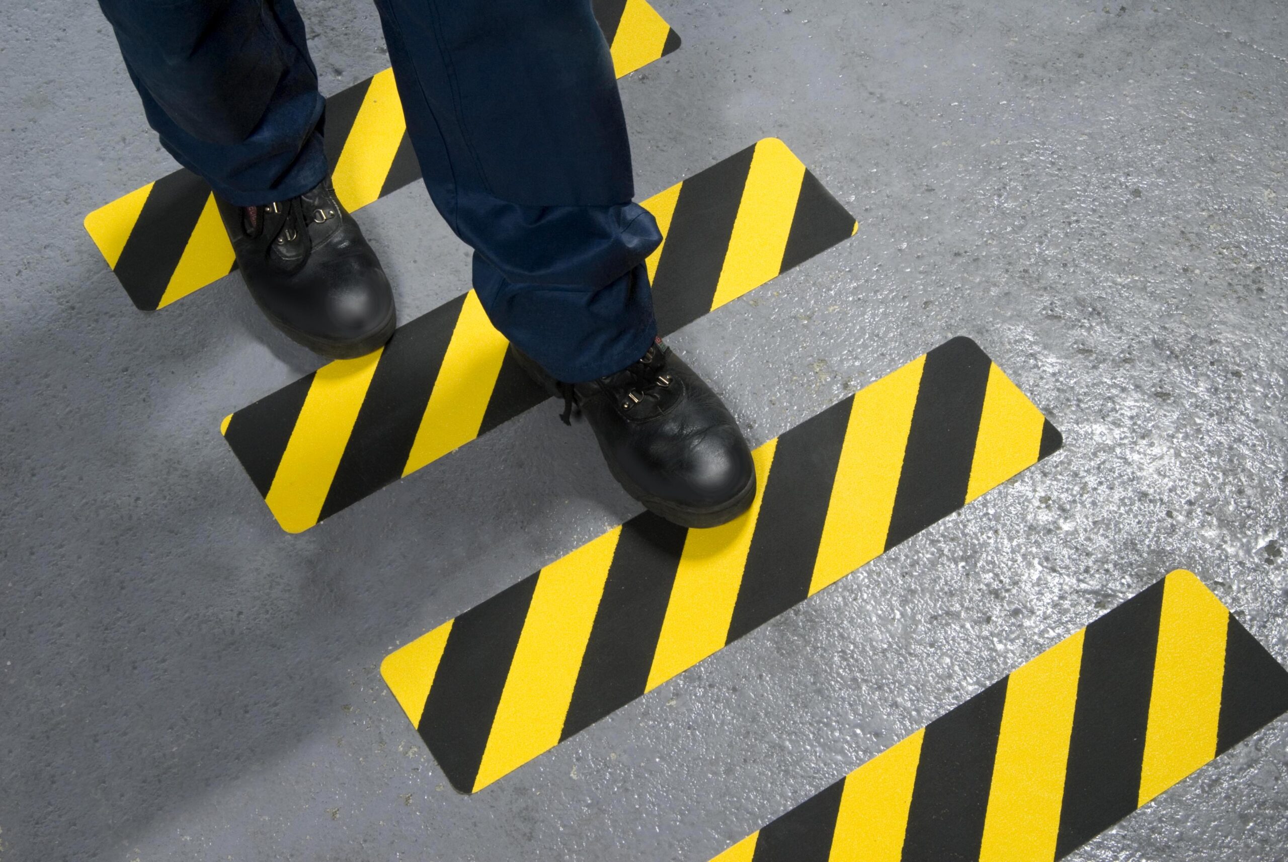 Medewerker staat met werkschoenen op geel-zwarte antislip vloerstrips met diagonale strepen op een betonnen vloer.