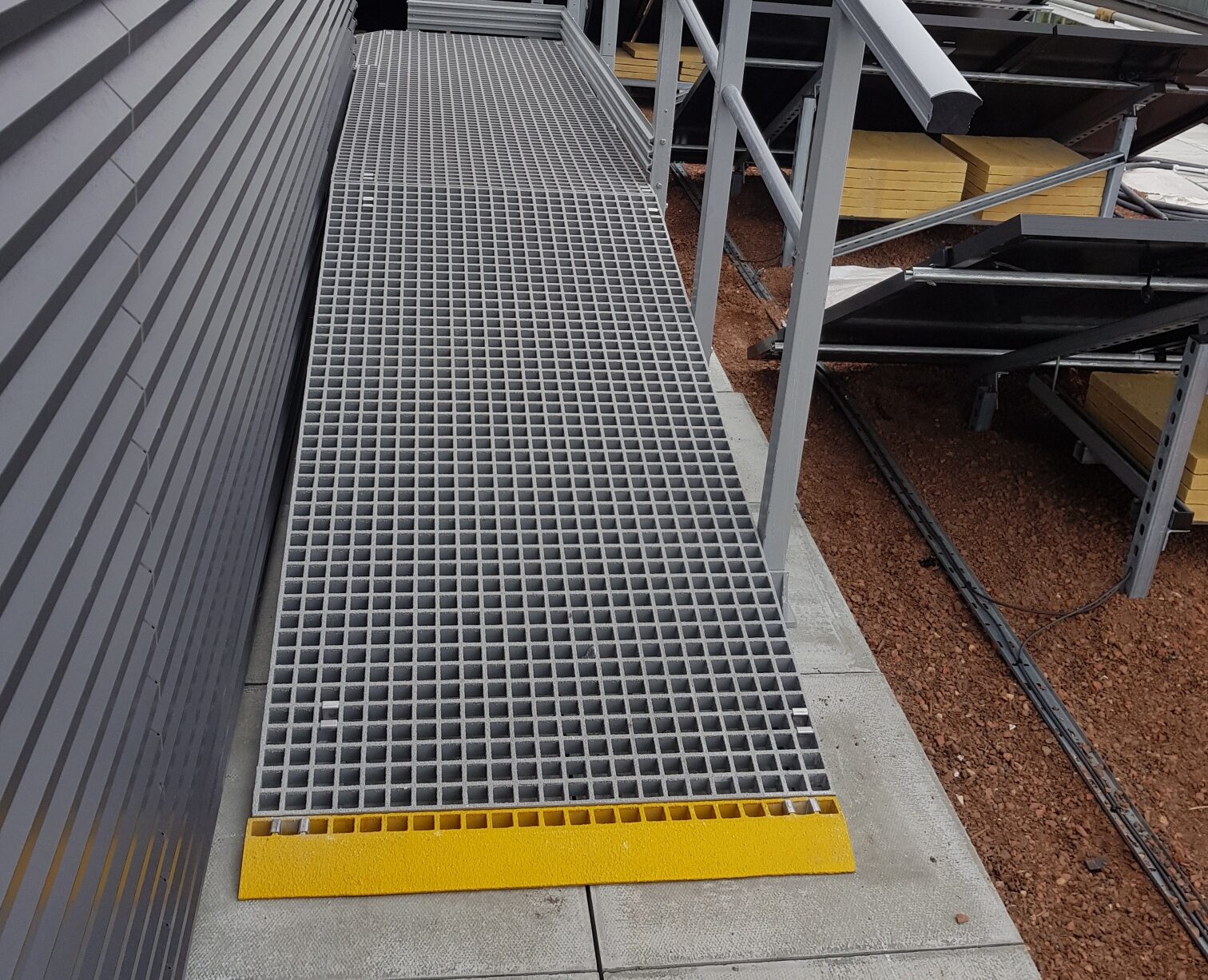 Gegalvaniseerde stalen loopbrug met roosteroppervlak en gele GRP antislip trapneus, gelegen op een dak met zicht op stedelijke omgeving