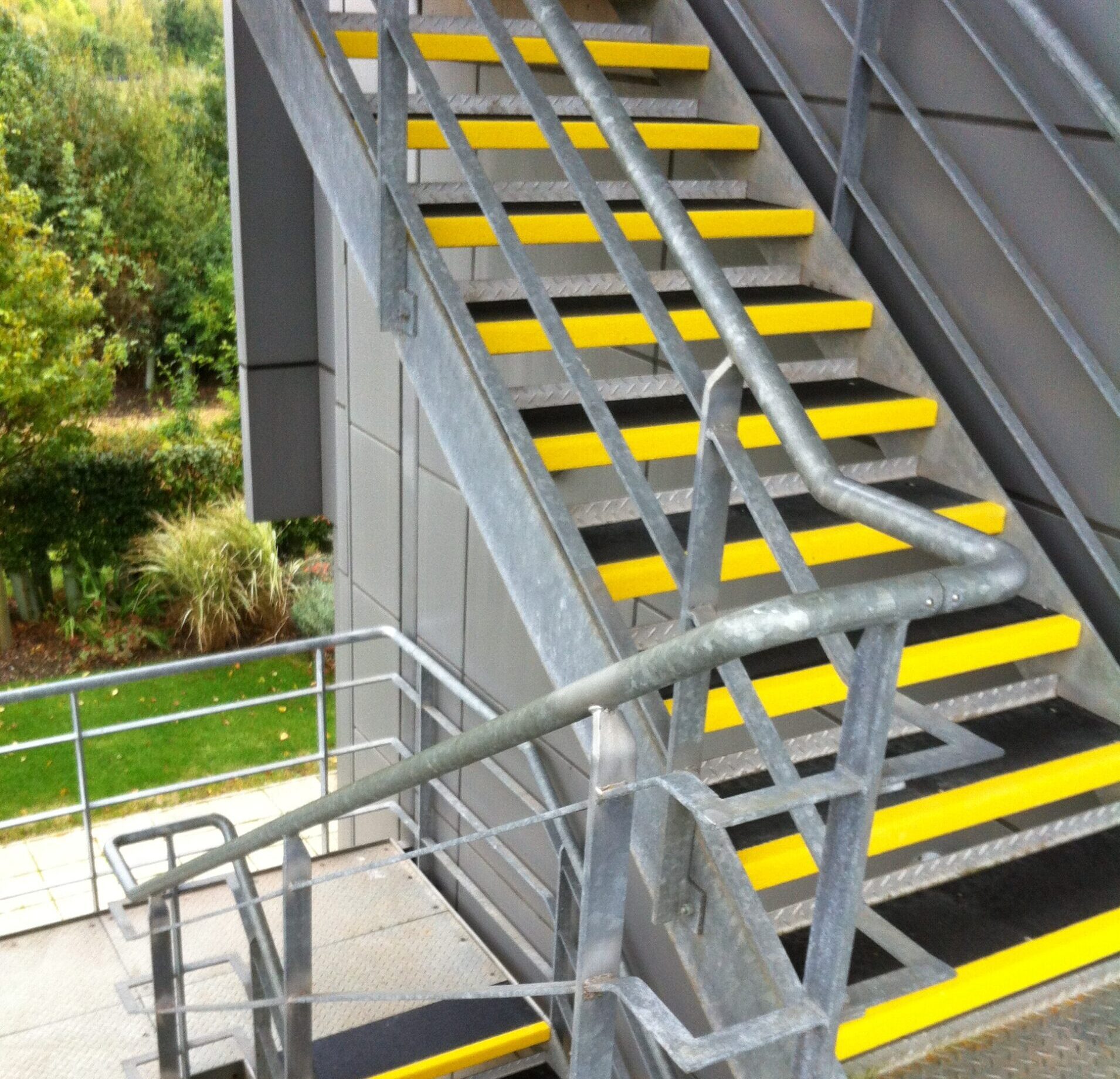 Stalen buitentrap met gele antislip GRP trapbedekkingen op elke trede, met metalen leuning en groene achtergrond.