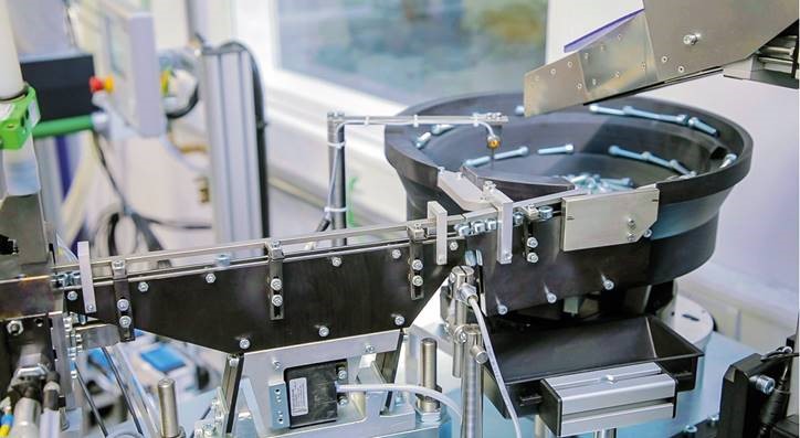 Industriële deprag bowlfeeder met ronde trilschaal gevuld met schroeven, verbonden met een geleidingsbaan in een geautomatiseerde productieomgeving.