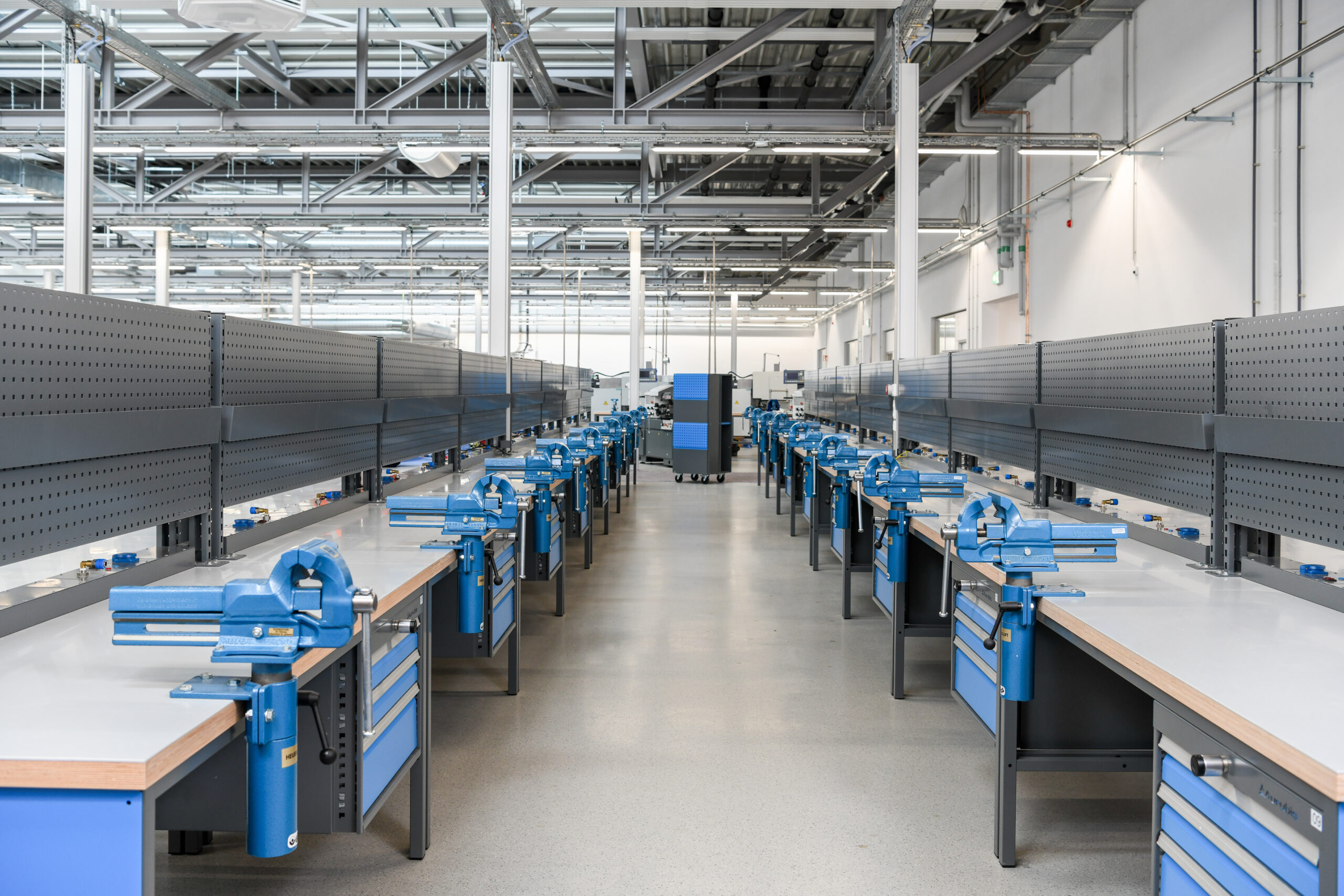 Lange rijen professionele werktafels met blauwe bankschroeven, ladekasten en grijze geperforeerde panelen in een ruime industriële werkplaats - Longues rangées de tables de travail professionnelles avec étaux bleus, caissons à tiroirs et panneaux perforés gris dans un vaste atelier industriel