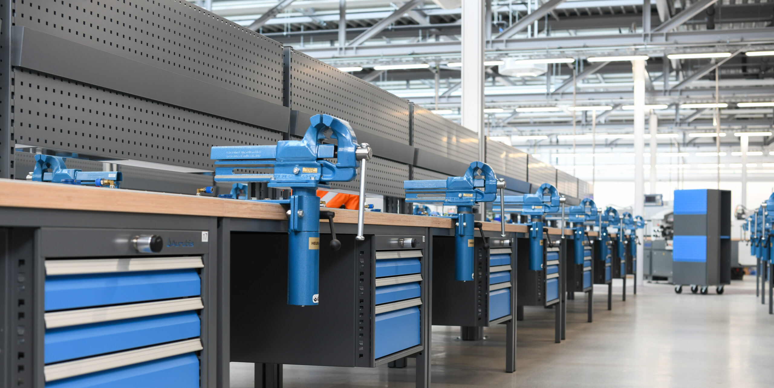 Rij blauwe werkbanken met ladekasten en perfopanelen in een industriële productiehal