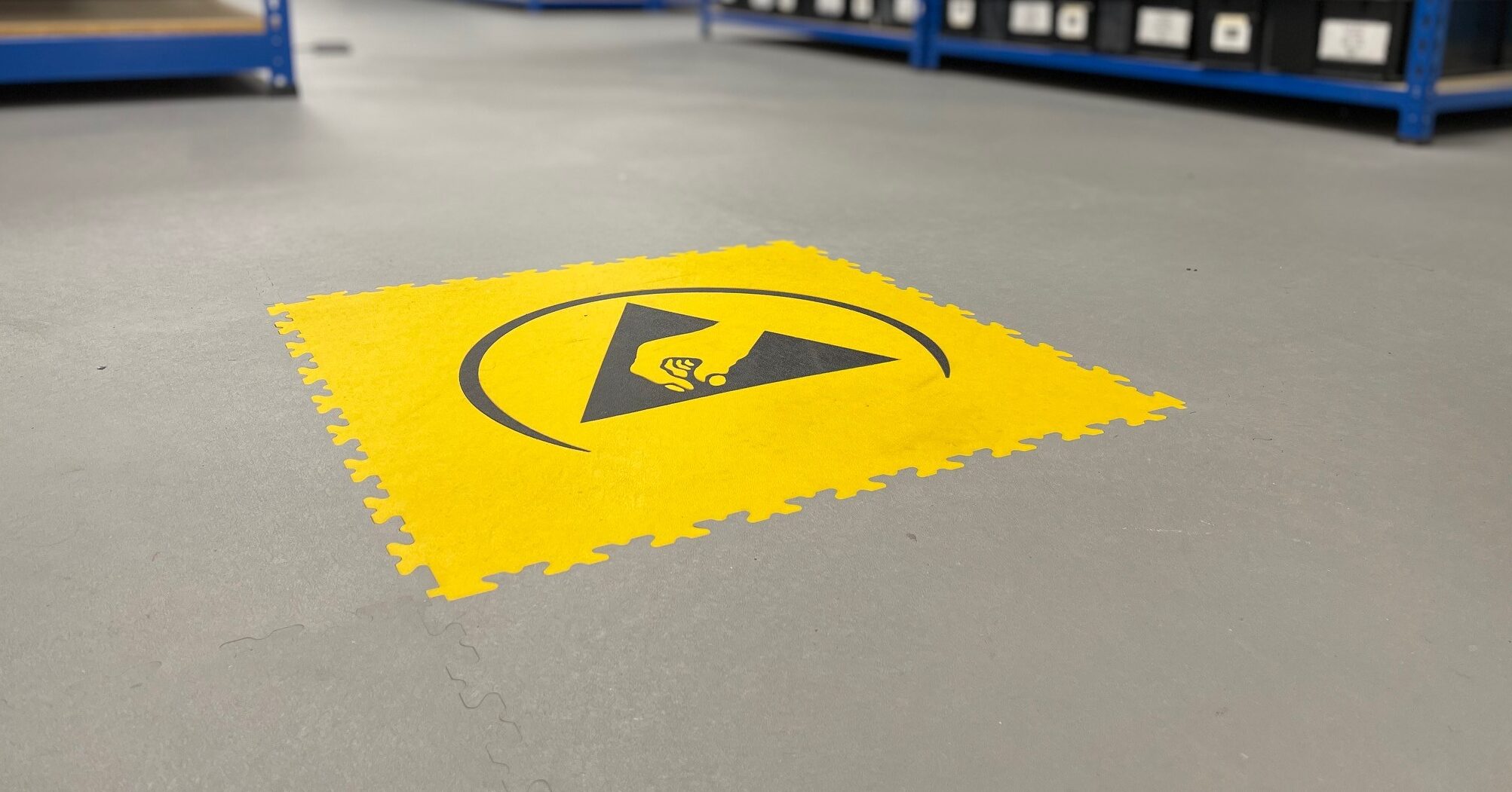 Gele ESD vloertegel met zwart antistatisch waarschuwingssymbool op een grijze industriële vloer, met blauwe stellingkasten op de achtergrond.