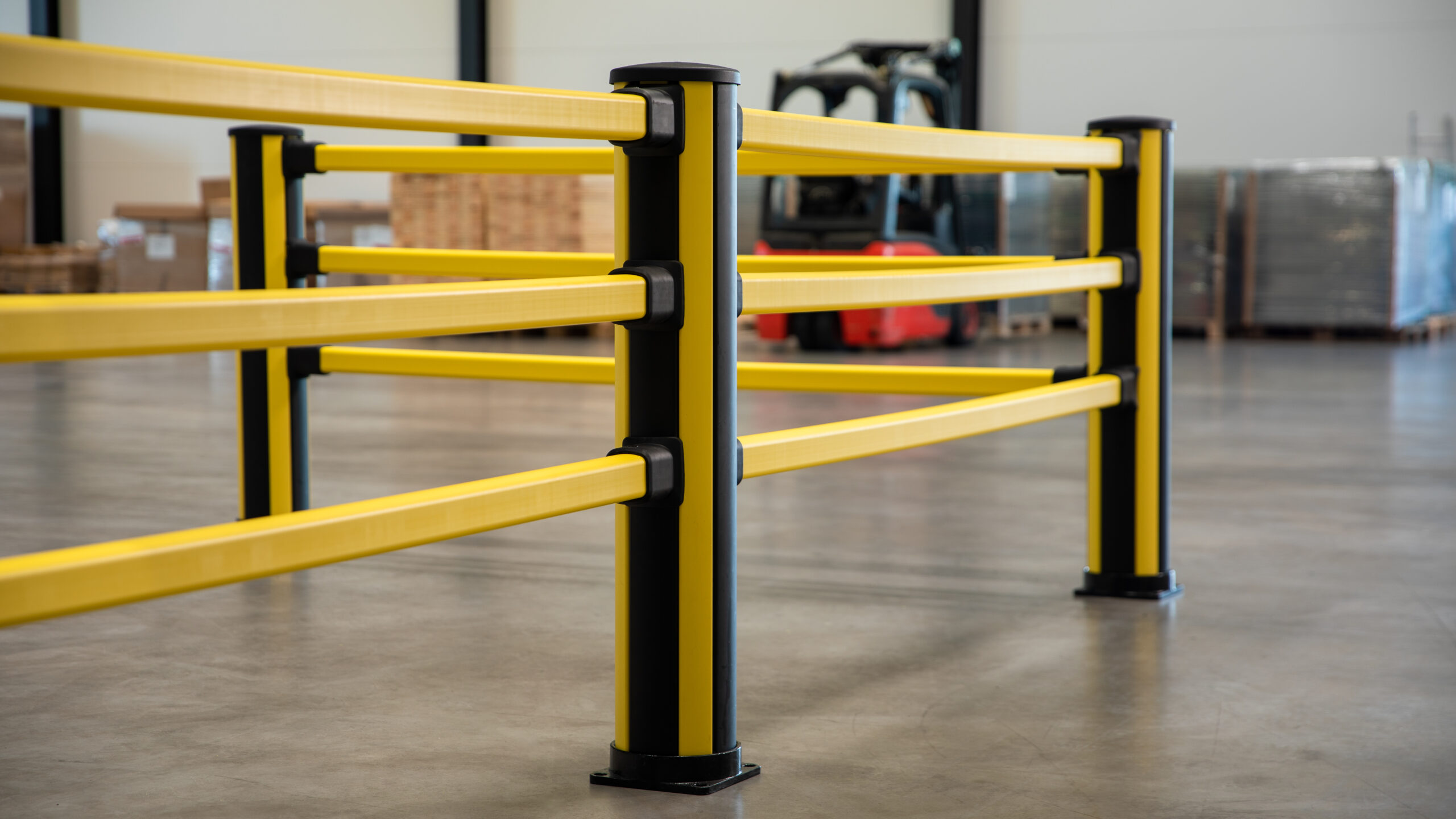 Close-up van een gele drievoudige verkeersbarrière met zwarte vierkante palen op een betonnen magazijnvloer, met op de achtergrond een rode vorkheftruck en pallets