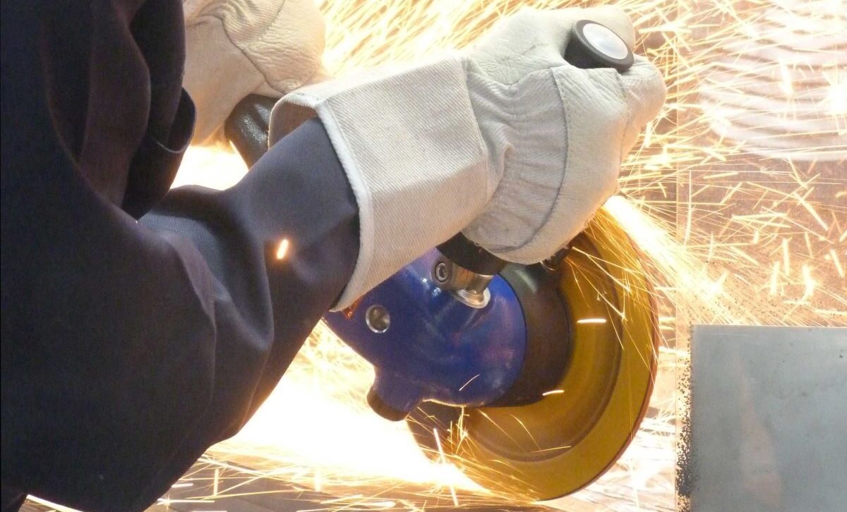 Medewerker met beschermende handschoenen gebruikt een blauwe Deprag pneumatische haakse slijpmachine op een metalen werkstuk, met vonken rondom.