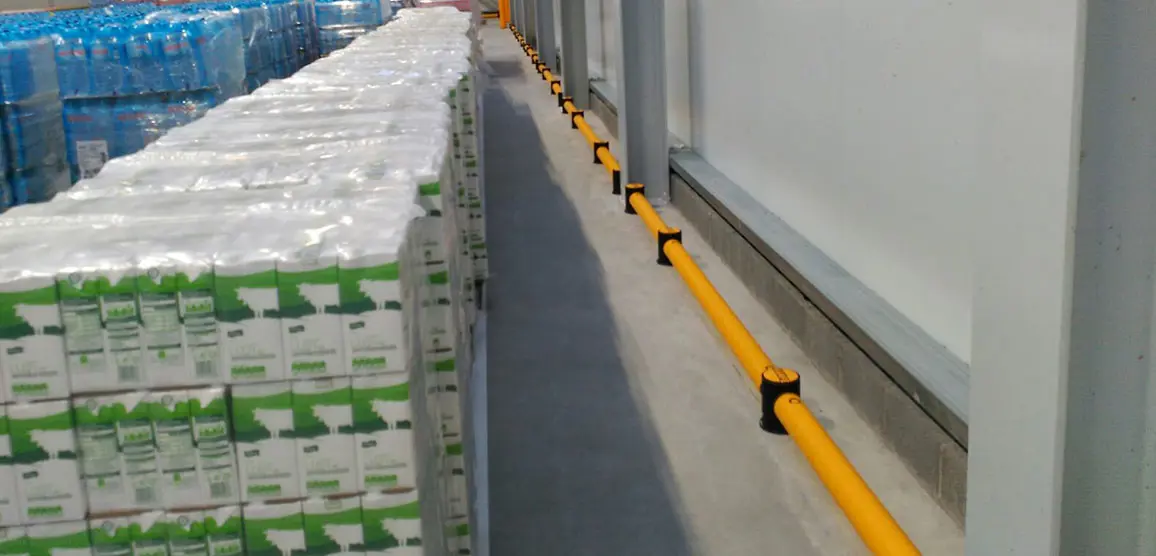 Rij geel-zwarte microbarrières bevestigd langs een witte magazijnwand naast opgeslagen pallets met goederen.