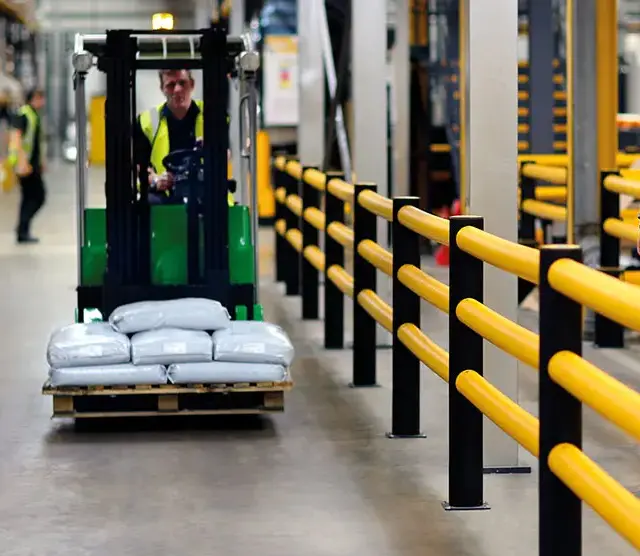 Groene heftruck rijdt langs een rij geel-zwarte verkeersbarrières die een rijstrook afscheiden van de voetgangerszone in een magazijn.