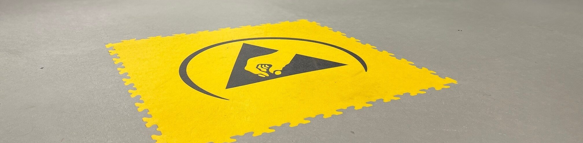 Gele ESD-vloertegel met het internationaal erkend ESD-symbool, aangebracht op een grijze industriële vloer ter aanduiding van een elektrostatisch beschermde werkzone. Carreau jaune ESD, marquant avec l’icône ESD reconnue internationalement, placé sur un sol industriel gris pour indiquer clairement une zone protégée contre les décharges électrostatiques.