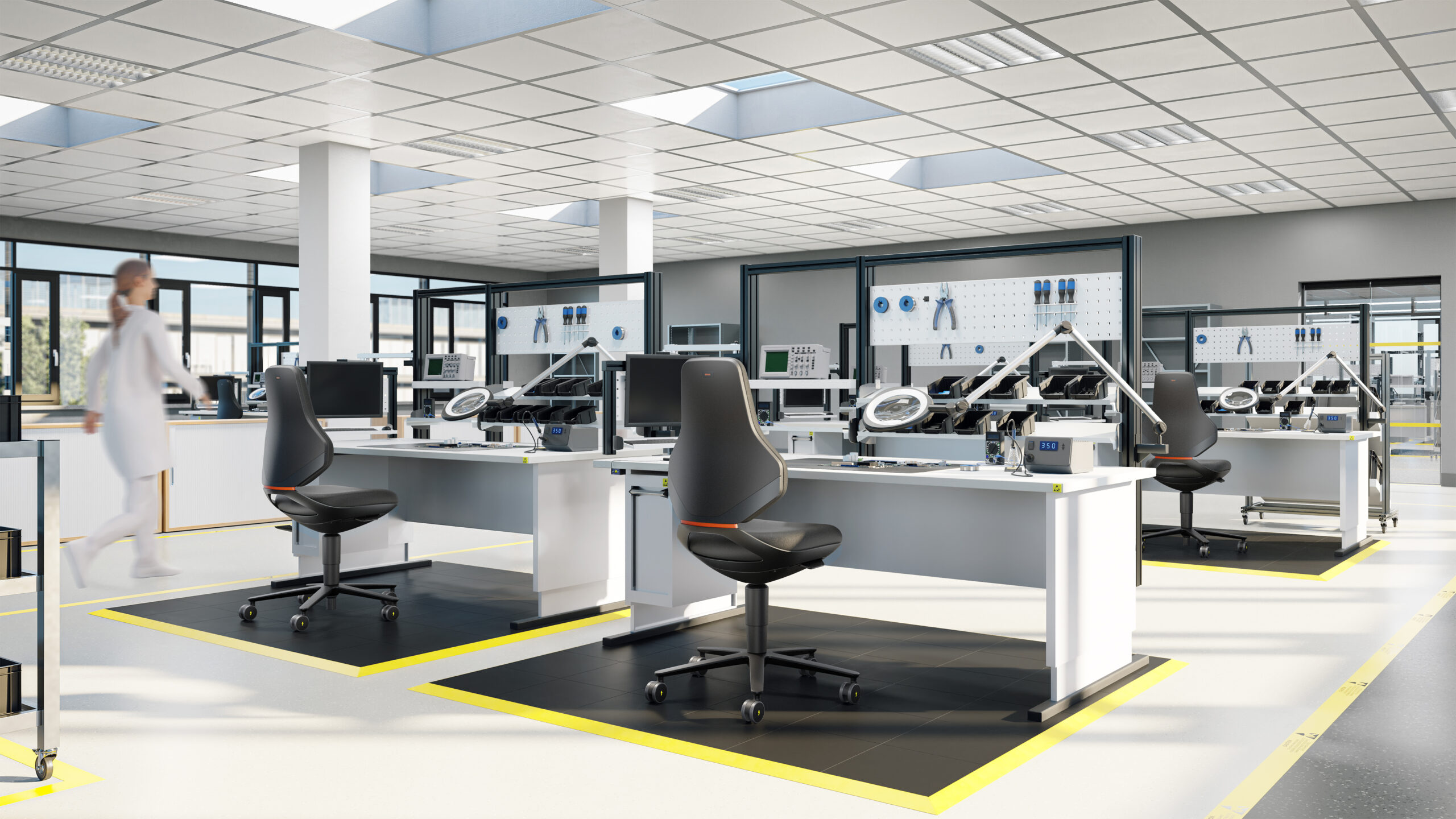 Een ESD‑werkzone met witte, in hoogte verstelbare ESD‑tafels en bijpassende ESD‑stoelen. Onder elke werkpost ligt een zwarte ESD‑mat, duidelijk omlijnd met gele markeringen. Op de achtergrond werken medewerkers in een professionele elektronica‑omgeving. Une zone de travail ESD avec des tables ESD blanches réglables en hauteur et des chaises ESD assorties. Sous chaque poste de travail se trouve un tapis ESD noir, clairement délimité par des marquages jaunes. En arrière-plan, des employés travaillent dans un environnement électronique professionnel.