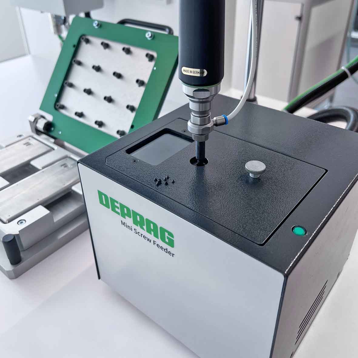 Deprag Mini Screw Feeder op een werktafel met een schroefspindel die via de bovenzijde van het apparaat schroeven aanvoert, met op de achtergrond een groen montagejig met geplaatste schroeven