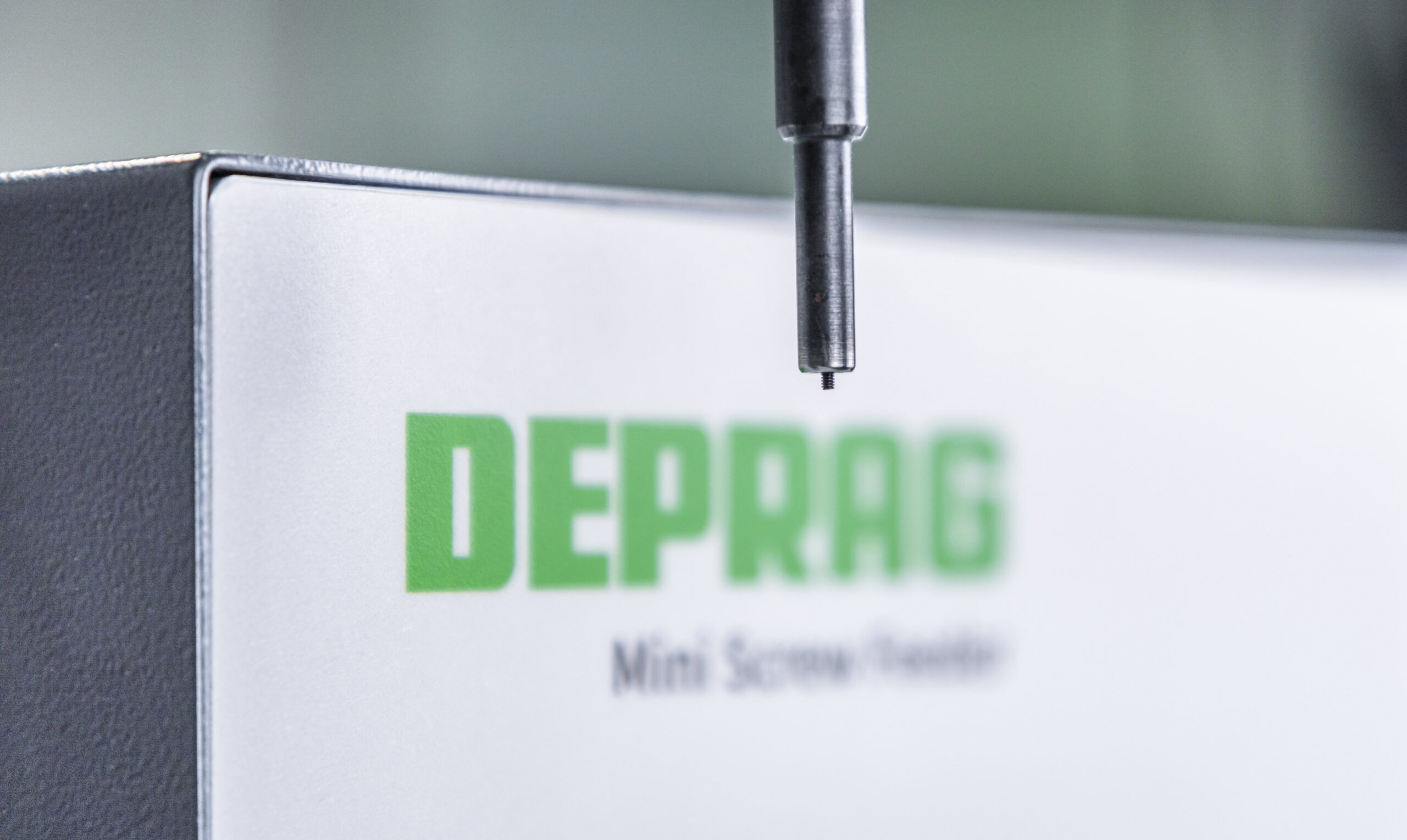 Close-up van een Deprag schroevendraaierbit met een minischroef aan de punt, voor een onscherpe achtergrond met het logo en de naam Deprag Mini Screw Feeder
