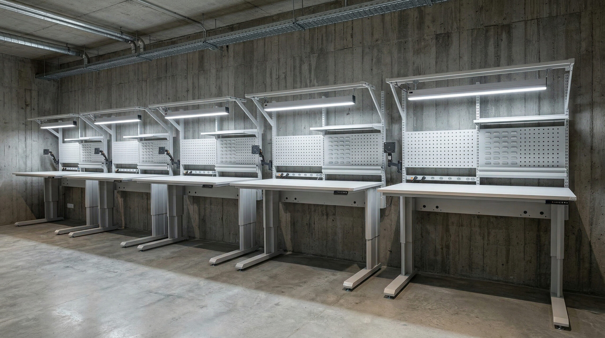 Vijf identieke elektrisch verstelbare werktafels met LED-verlichting en geperforeerde achterpanelen, opgesteld in een rij tegen een betonnen wand