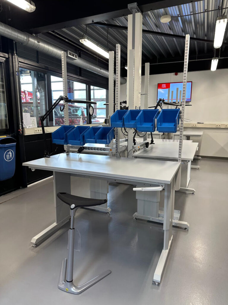 Rij in hoogte verstelbare ergonomische werktafels met witte werkbladen en witte bovenstellingen in een brandweeronderhoudswerkplaats, met een ergonomische zadelstoel op de voorgrond