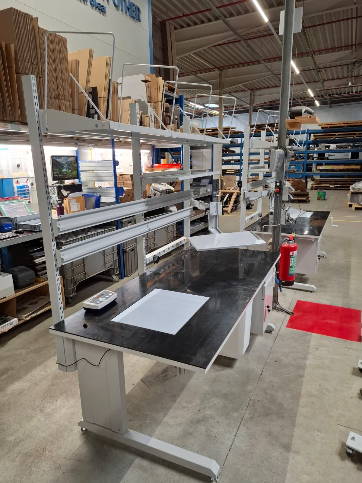 Schuin aanzicht van een industriële werktafel met zwart werkblad en aluminium bovenstelling met documenthouder, een weegschaal op het blad en een brandblusser ernaast