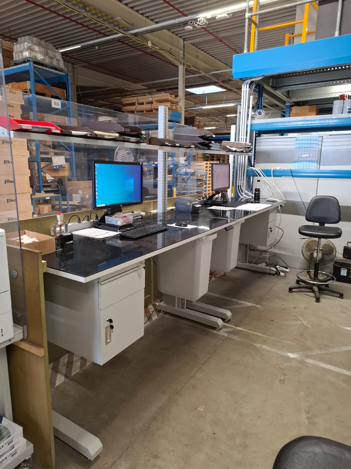 Twee industriële werktafels met zwart werkblad in gebruik, voorzien van computerschermen, toetsenborden, ladeblokken en een acrylaat spatscherm, met een bureaukruk naast de werkposten