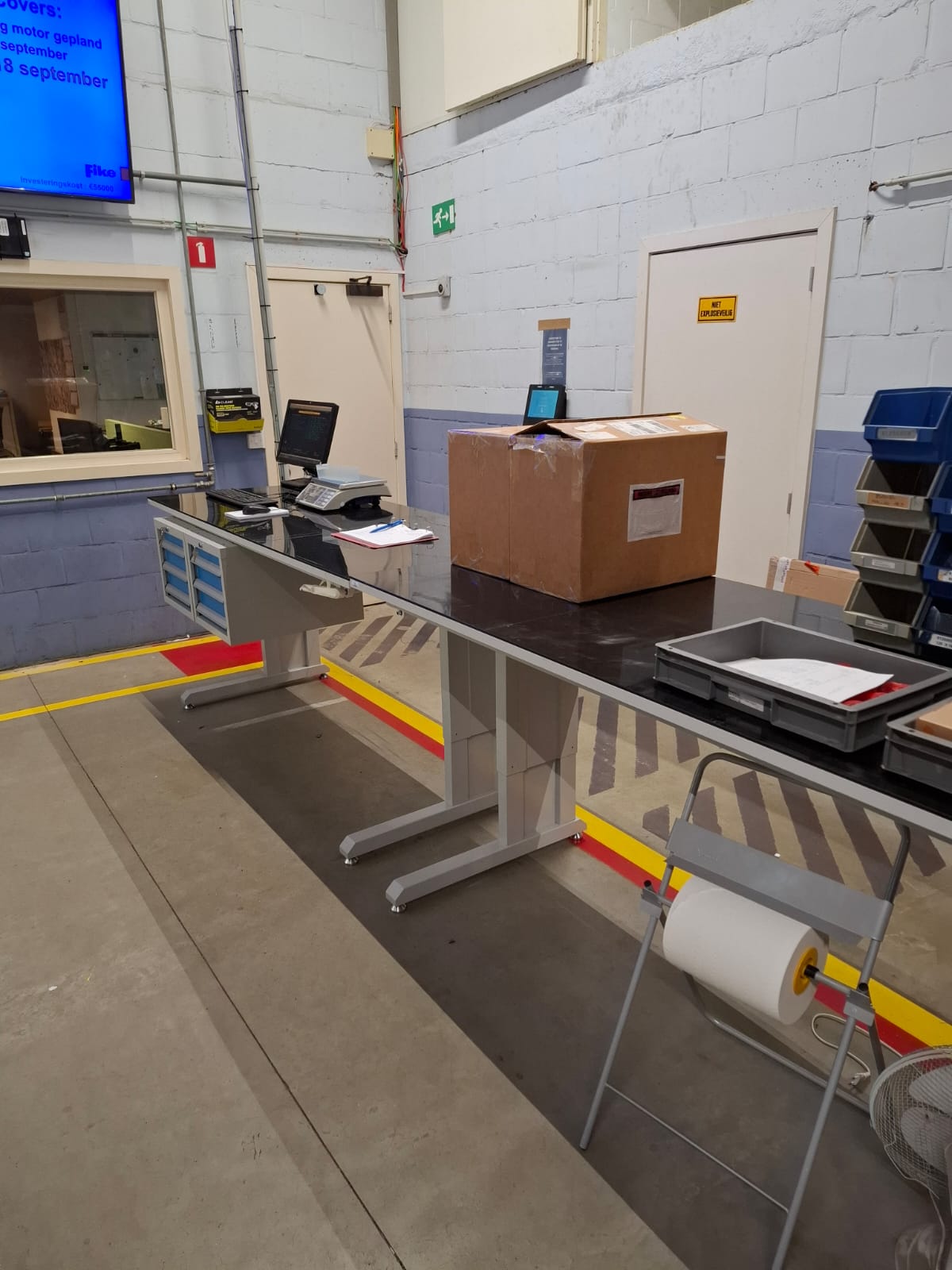 Industriële werktafel met zwart werkblad in gebruik in een productieomgeving, met een weegschaal, barcodescanner, documenthouder en een doos op het blad, en een papierrolhouder naast de tafel