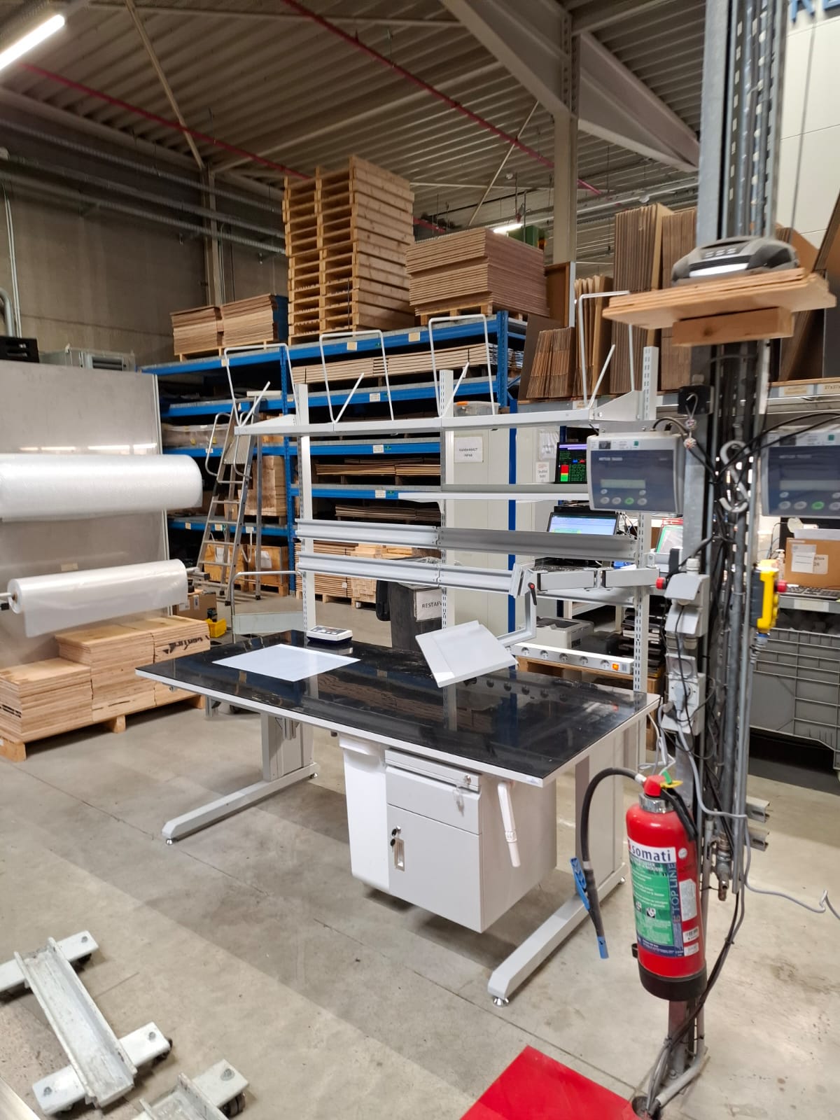 Zijaanzicht van een industriële werktafel met zwart werkblad, bovenstelling, folierolhouder links en weegapparatuur met display rechts, opgesteld in het AWP-atelier
