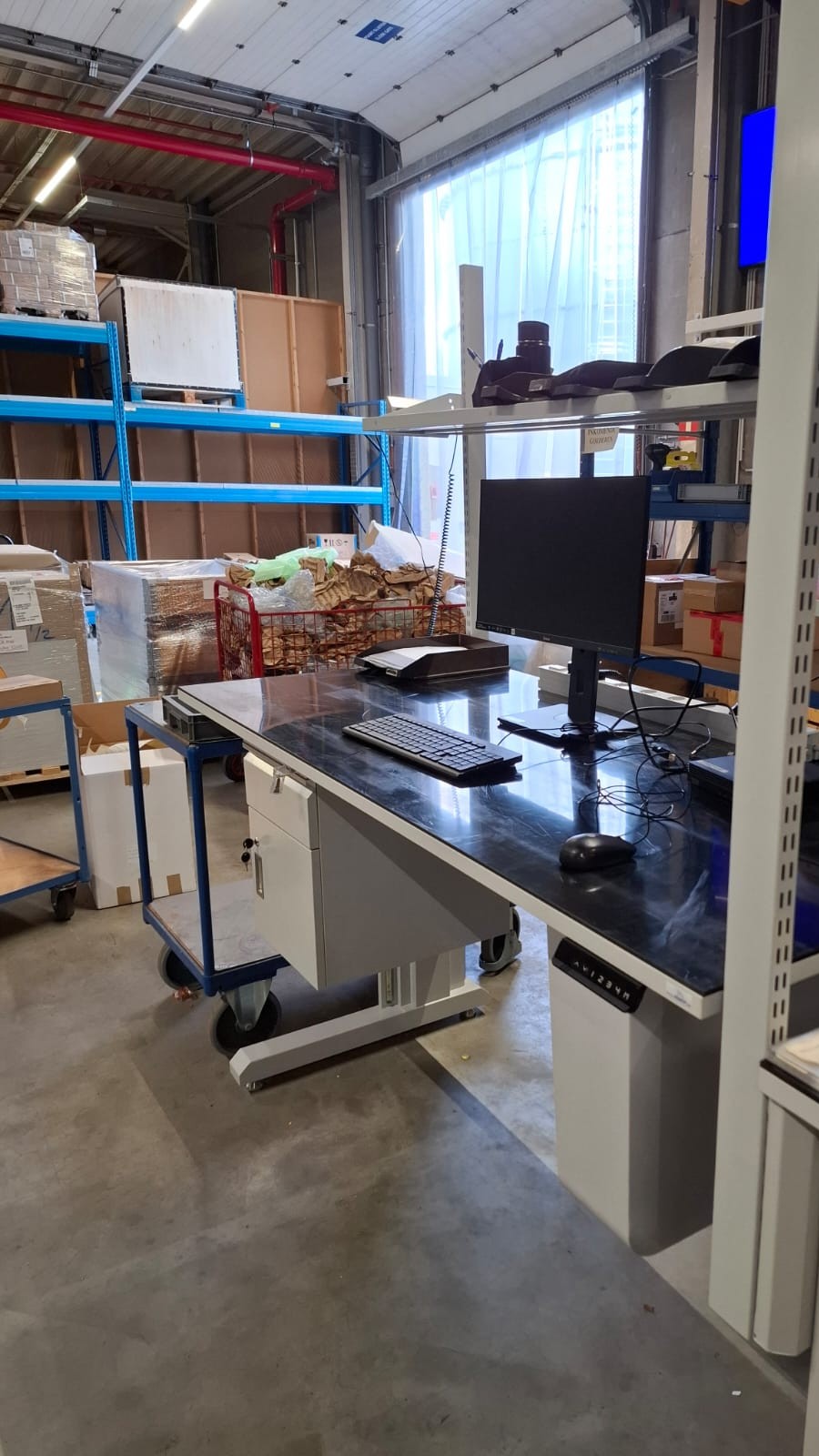 Enkele werkpost met zwart werkblad, monitor, toetsenbord en ladeblok in een magazijnomgeving, met een verrijdbare kar en stellingen op de achtergrond