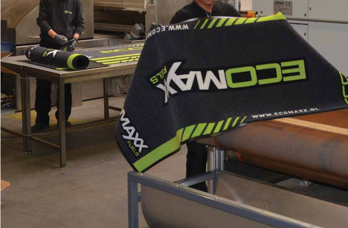 Twee medewerkers in een productieatelier die zwarte schoonveegmatten met geel-groene logobedrukking verwerken op een werkbank