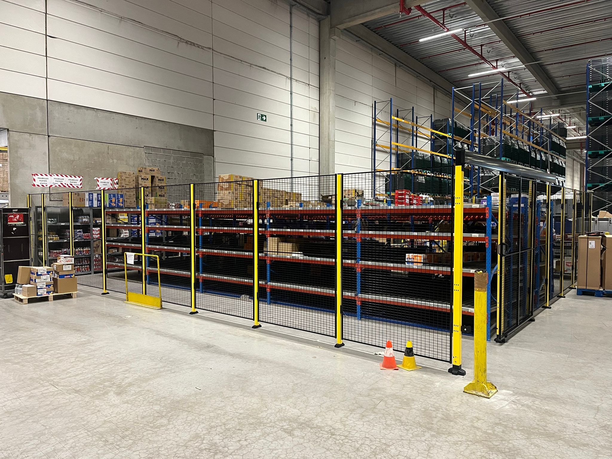 Lang hekwerk met gele staanders langs de voorzijde van magazijnstellingen vol dozen in een operationeel distributiecentrum, met oranje verkeerskegels op de vloer