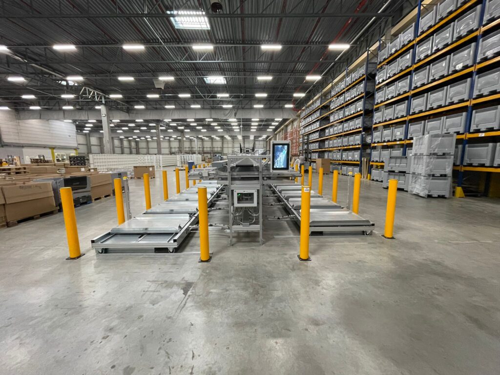 Totaaloverzicht van een sorteerzone in een groot distributiecentrum met gele veiligheidspaaltjes aan beide zijden van een geautomatiseerde sorteerinstallatie en hoge palletrekken vol grijze kratten op de achtergrond