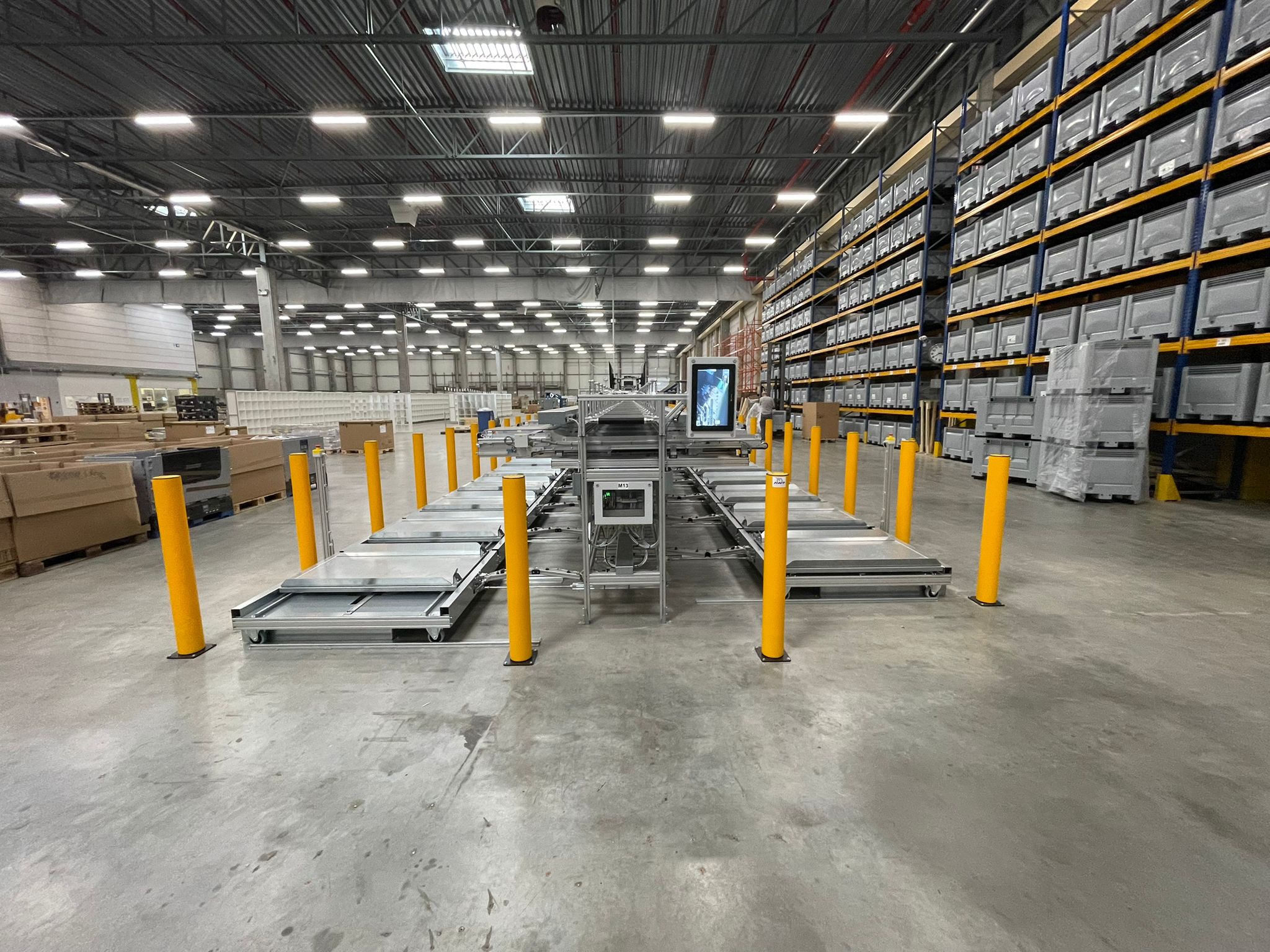 Totaaloverzicht van een sorteerzone in een groot distributiecentrum met gele veiligheidspaaltjes aan beide zijden van een geautomatiseerde sorteerinstallatie en hoge palletrekken vol grijze kratten op de achtergrond