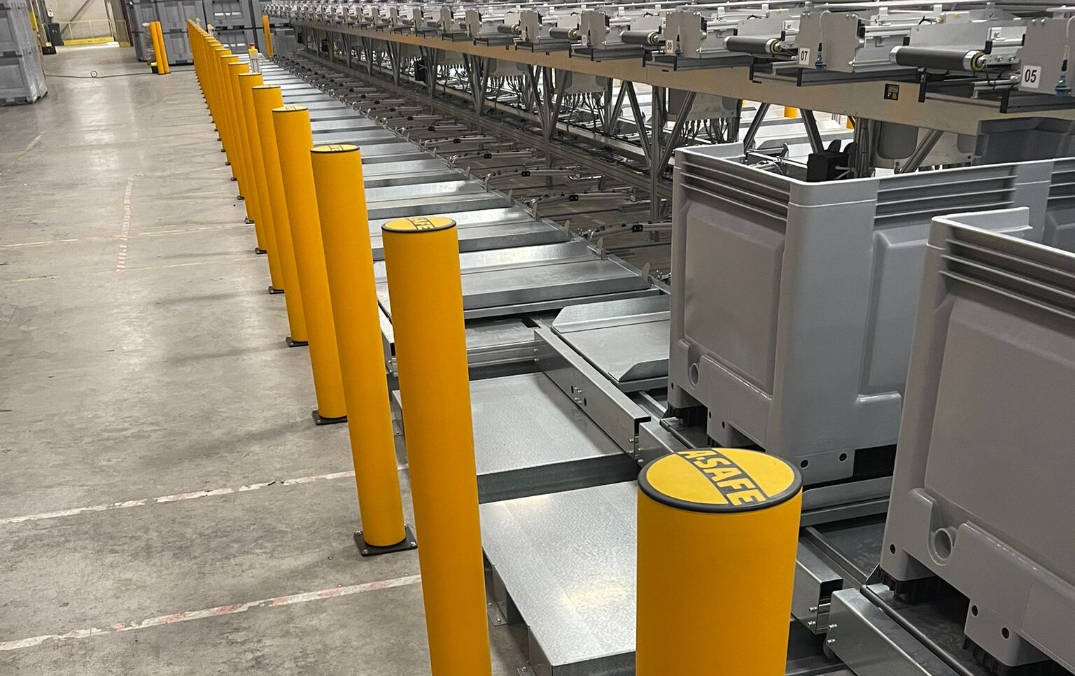 Lange rij gele ASAFE veiligheidspaaltjes langs een geautomatiseerde sorteerinstallatie met grijze transportbakken in een printdistributiecentrum