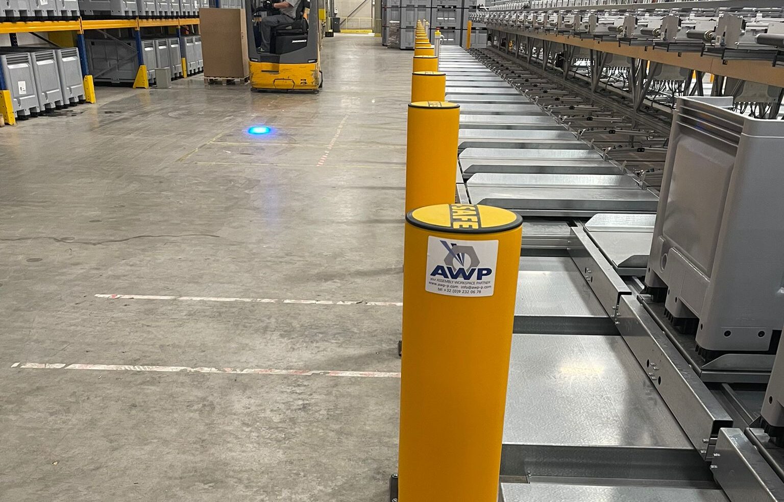 Close-up van een geel ASAFE veiligheidspaaltje met AWP-sticker langs een sorteerconveyor, met een rijdende heftruck en een blauw veiligheidslicht op de vloer op de achtergrond